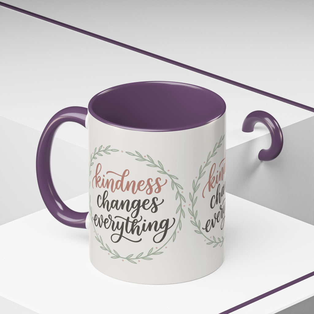 Inspirational ceramic accent coffee mug featuring hand-lettered “Kindness Changes Everything” typography surrounded by a delicate laurel wreath in soft sage and blush tones.