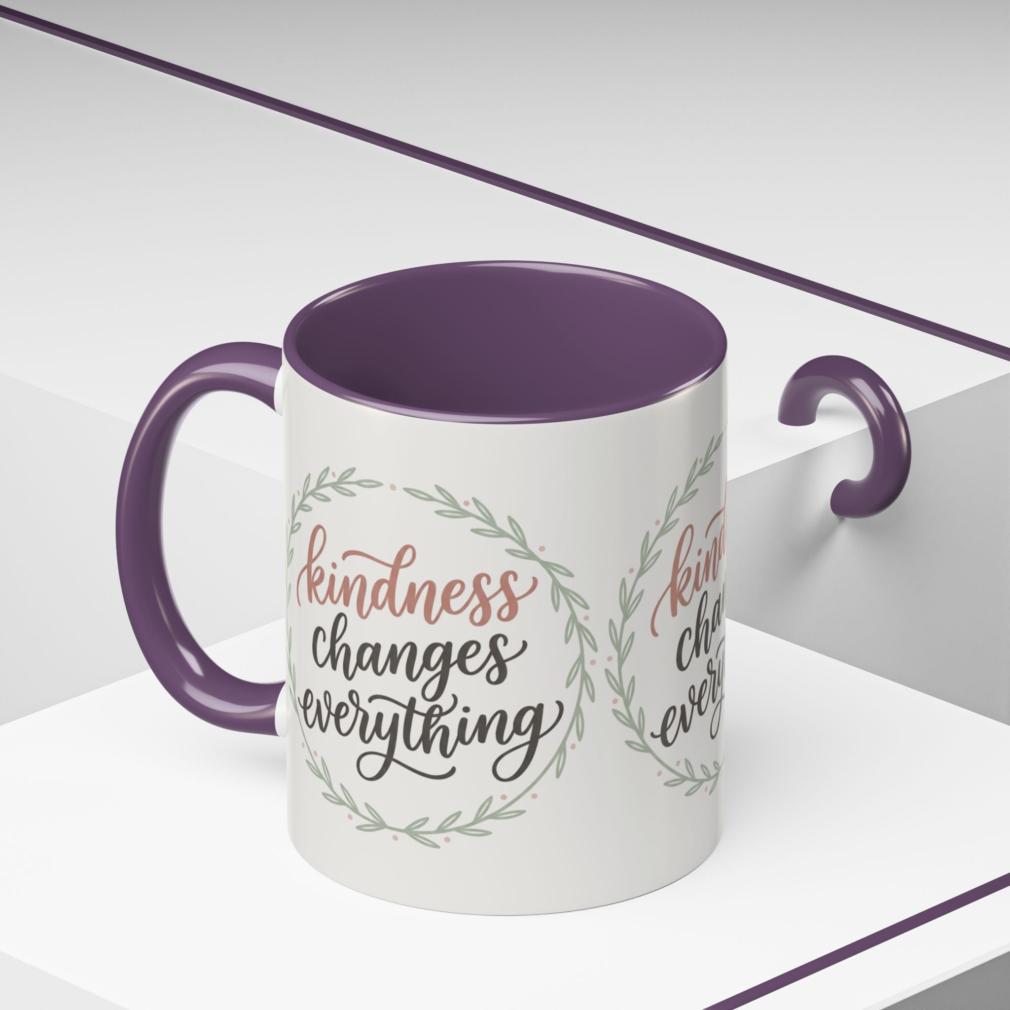 Inspirational ceramic accent coffee mug featuring hand-lettered “Kindness Changes Everything” typography surrounded by a delicate laurel wreath in soft sage and blush tones.