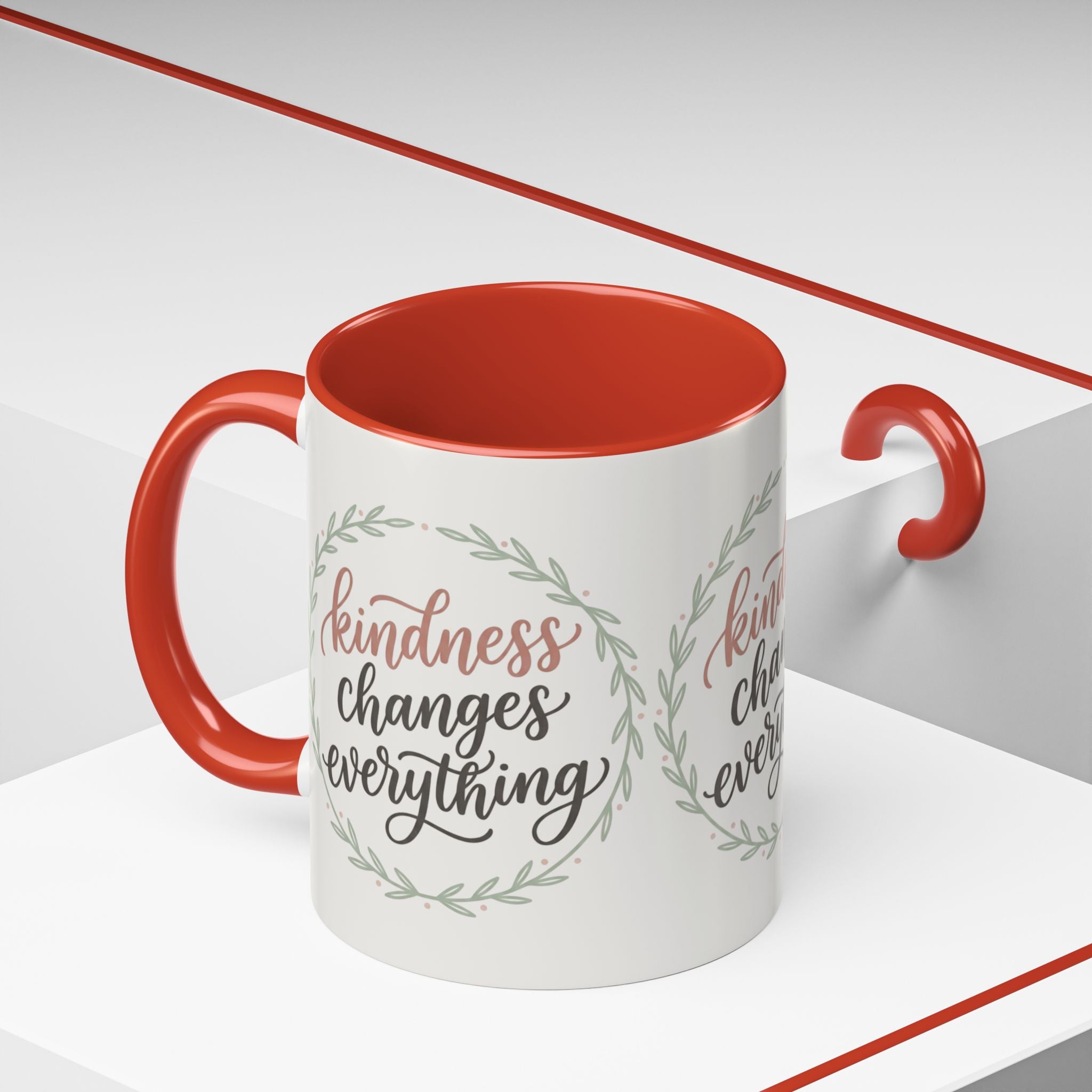 Inspirational ceramic accent coffee mug featuring hand-lettered “Kindness Changes Everything” typography surrounded by a delicate laurel wreath in soft sage and blush tones.