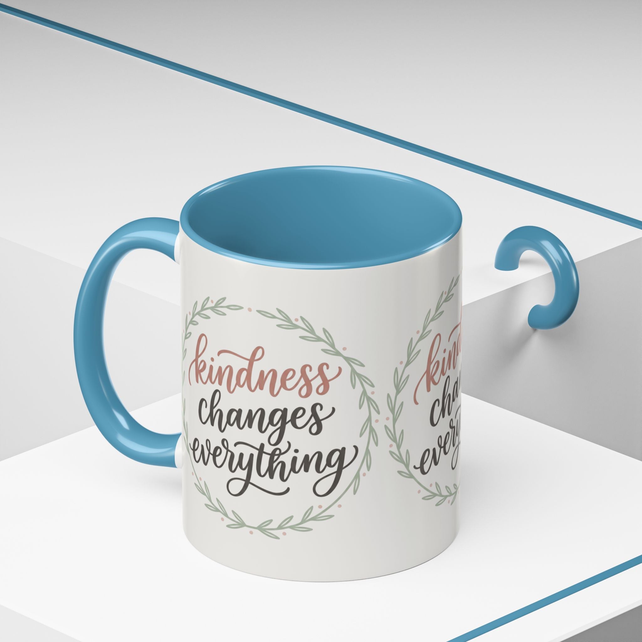 Inspirational ceramic accent coffee mug featuring hand-lettered “Kindness Changes Everything” typography surrounded by a delicate laurel wreath in soft sage and blush tones.