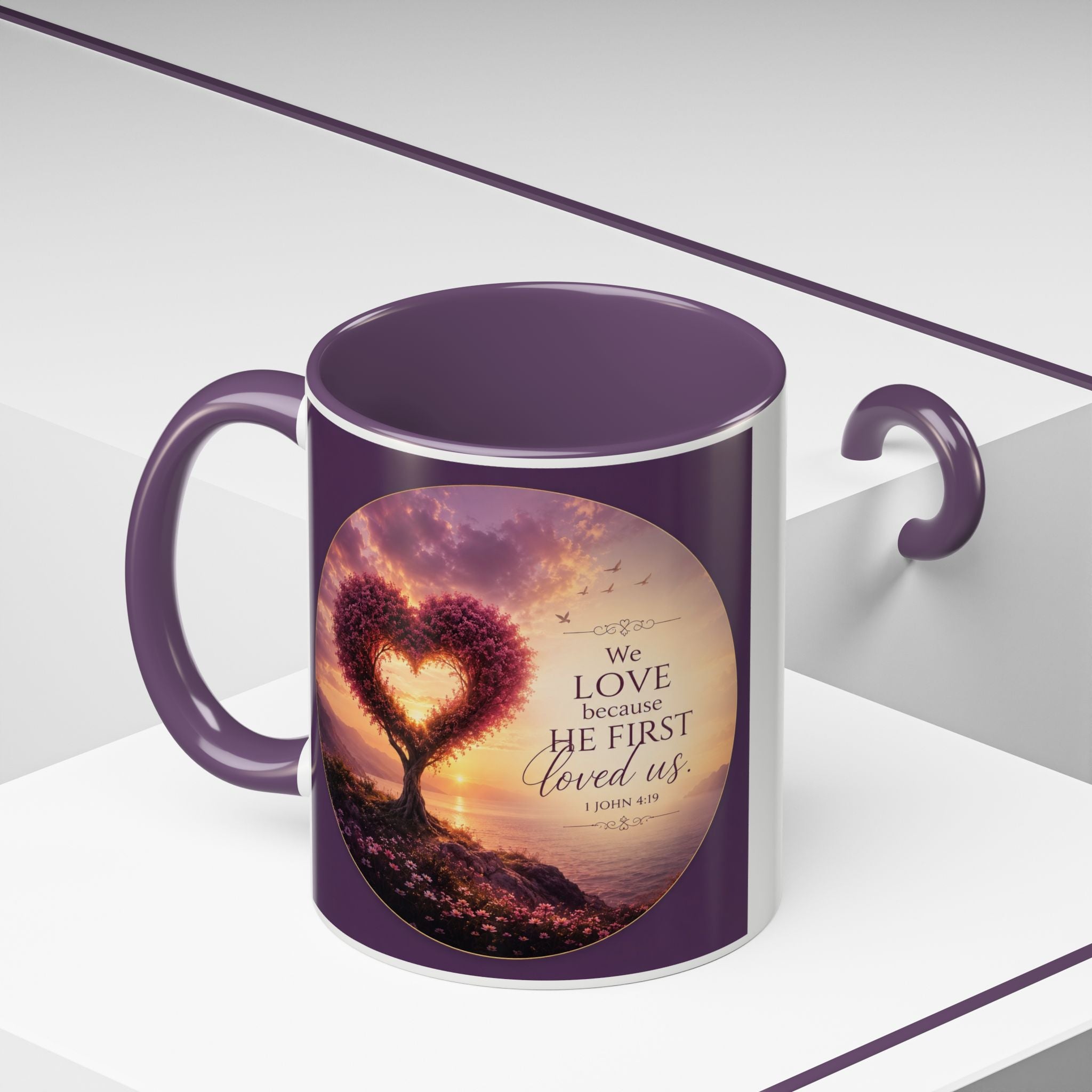 Inspirational Christian coffee mug featuring a glowing heart-shaped tree at sunset with the scripture “We Love Because He First Loved Us” from 1 John 4:19 and contrasting black handle and interior.