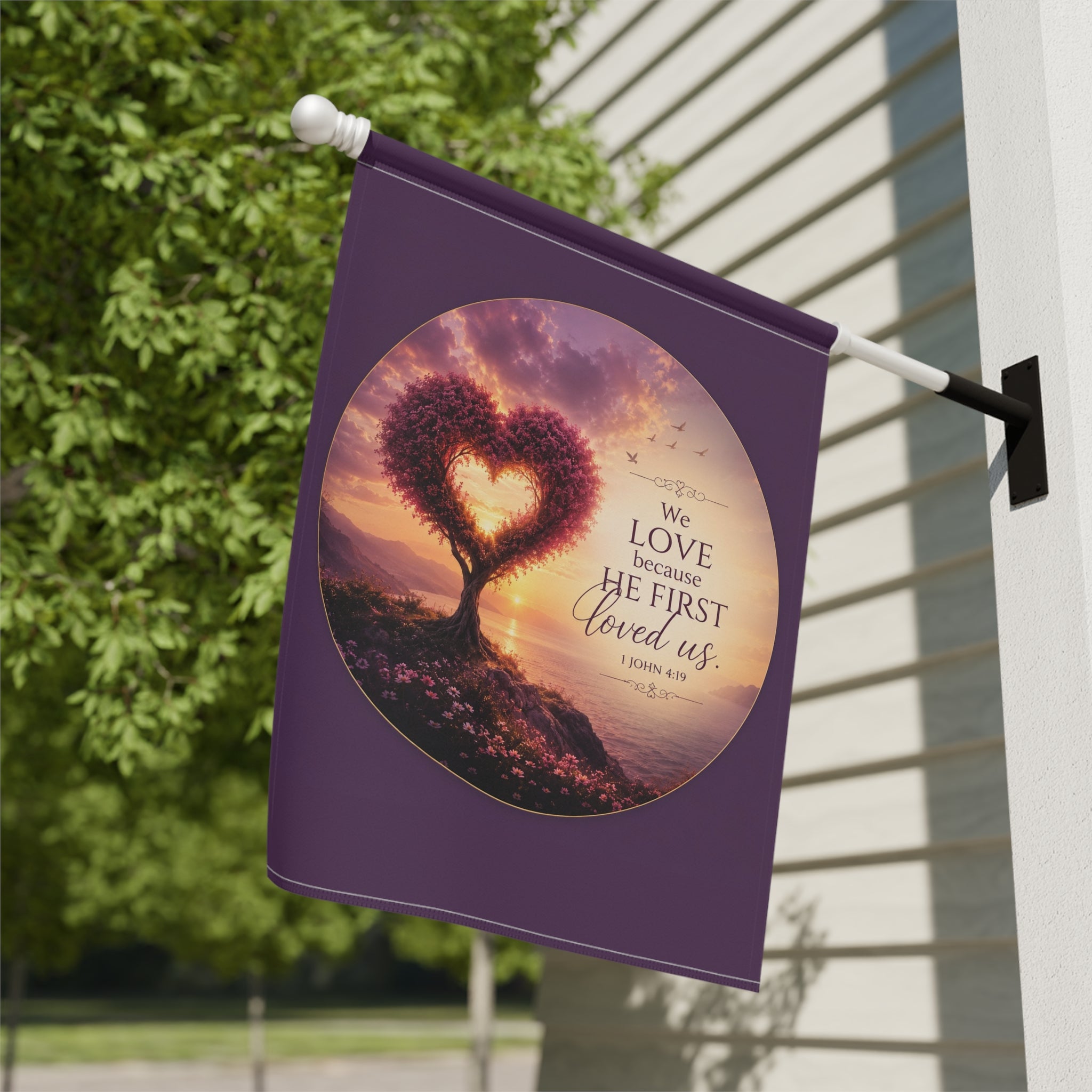 Inspirational Christian garden flag featuring a heart-shaped tree at sunset with the scripture “We Love Because He First Loved Us” in warm glowing tones.