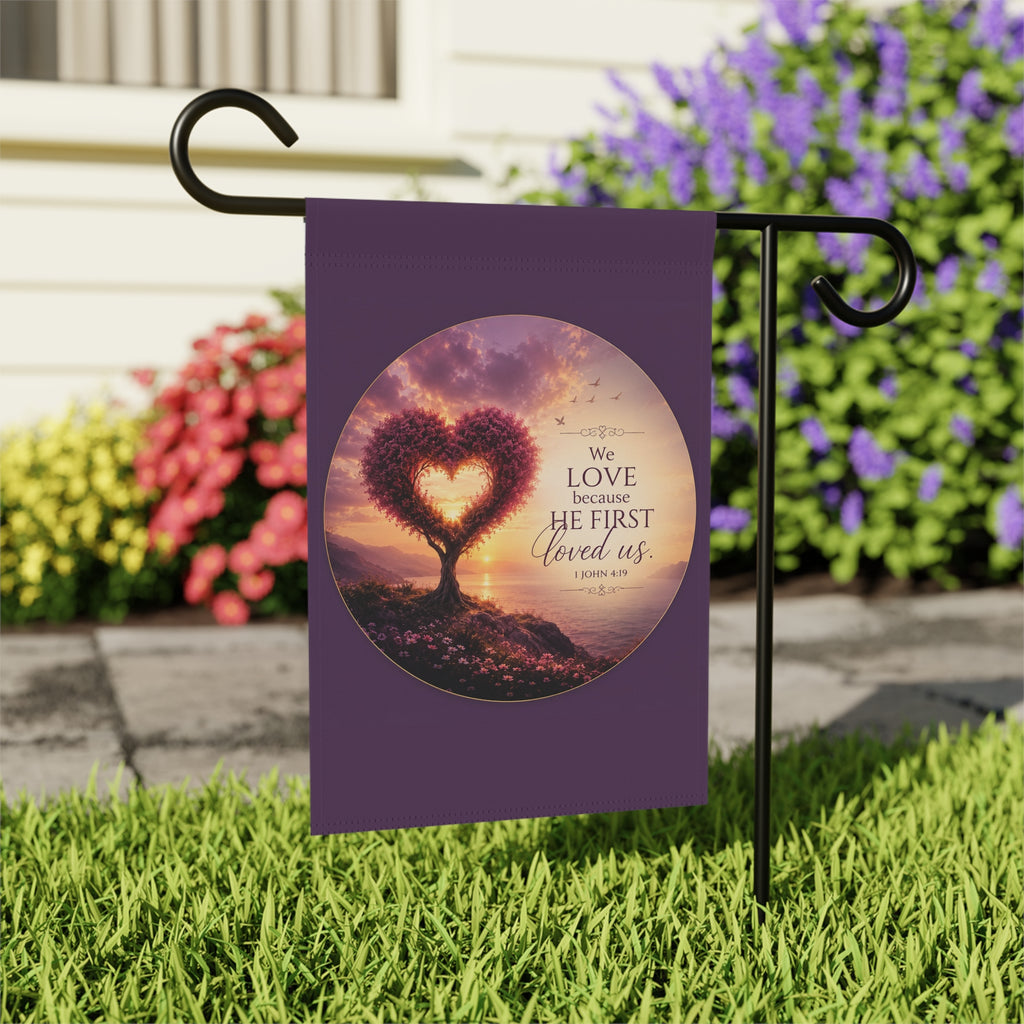 Inspirational Christian garden flag featuring a heart-shaped tree at sunset with the scripture “We Love Because He First Loved Us” in warm glowing tones.