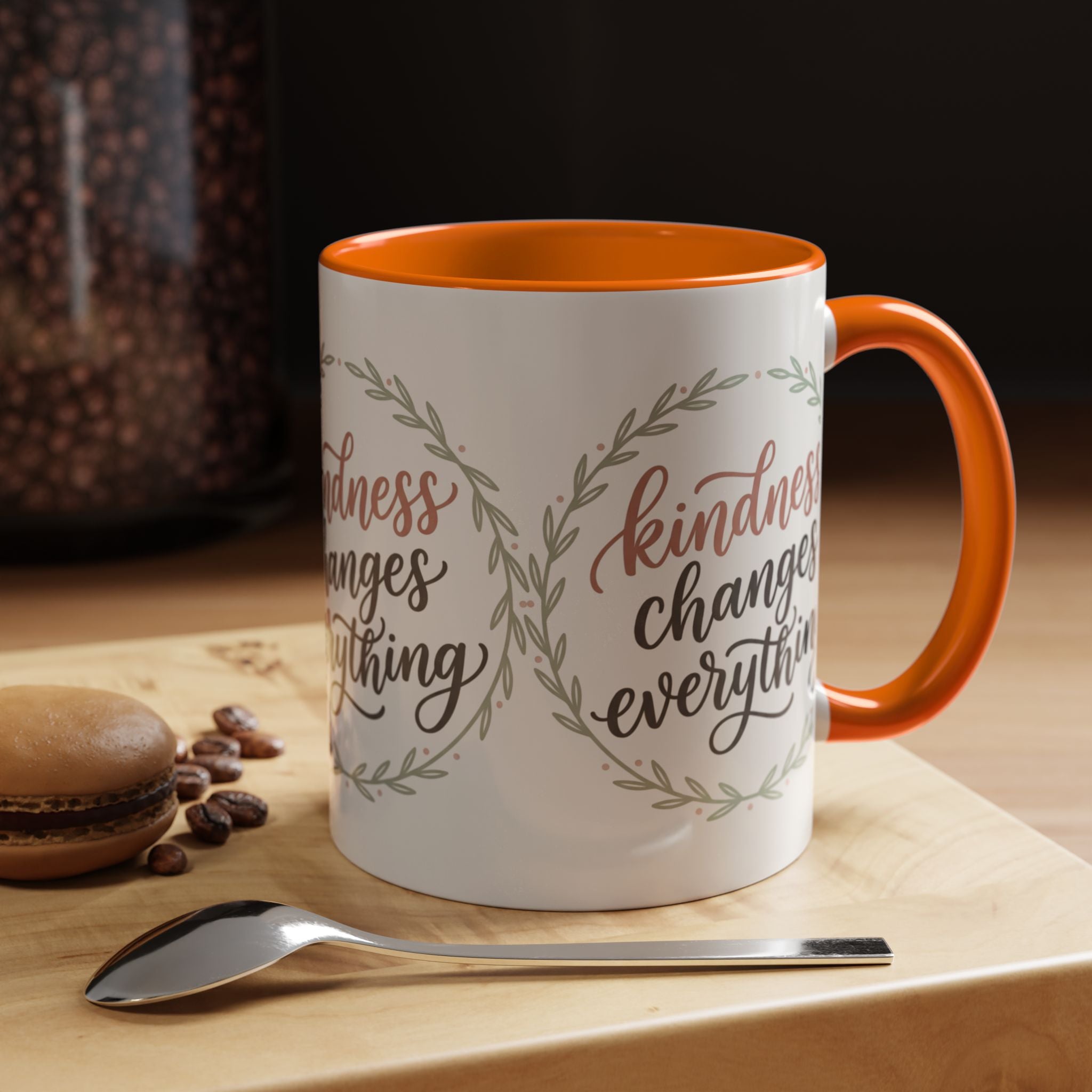 Inspirational ceramic accent coffee mug featuring hand-lettered “Kindness Changes Everything” typography surrounded by a delicate laurel wreath in soft sage and blush tones.