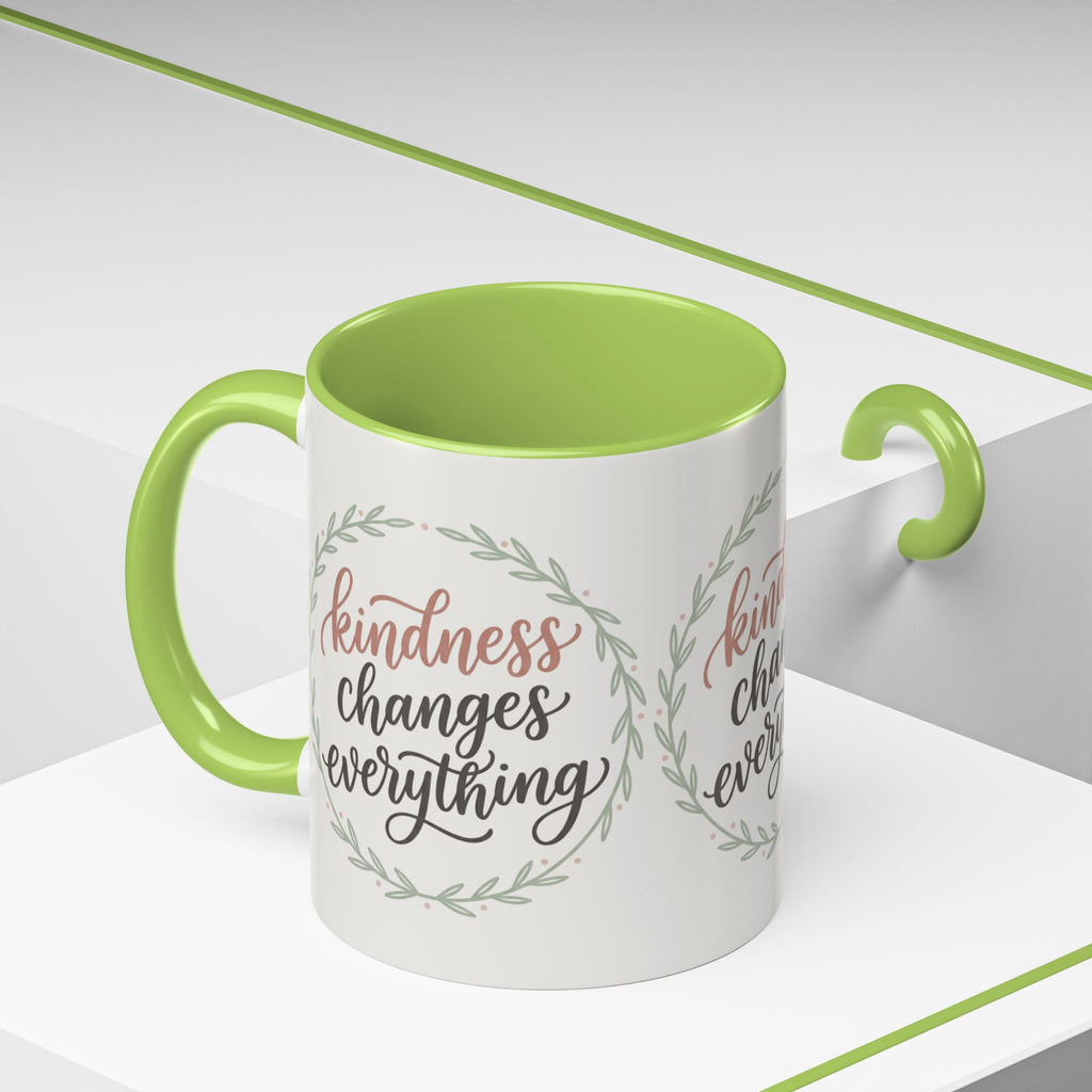 Inspirational ceramic accent coffee mug featuring hand-lettered “Kindness Changes Everything” typography surrounded by a delicate laurel wreath in soft sage and blush tones.
