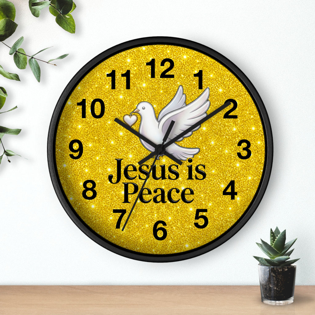 A round wall clock with a black frame and a glittery gold face, featuring a white dove, black numerals, and the words 'Jesus is Peace' in black text. The clock is shown mounted on a white wall above a light wood surface, with small green plants in the corners.