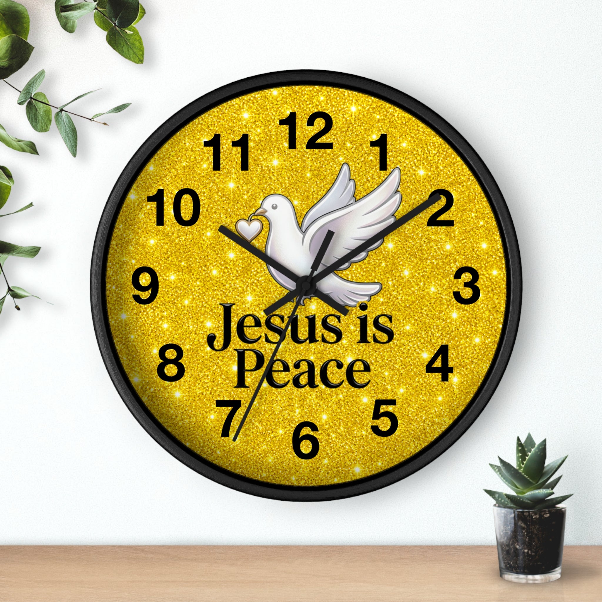 A round wall clock with a black frame and a glittery gold face, featuring a white dove, black numerals, and the words 'Jesus is Peace' in black text. The clock is shown mounted on a white wall above a light wood surface, with small green plants in the corners.