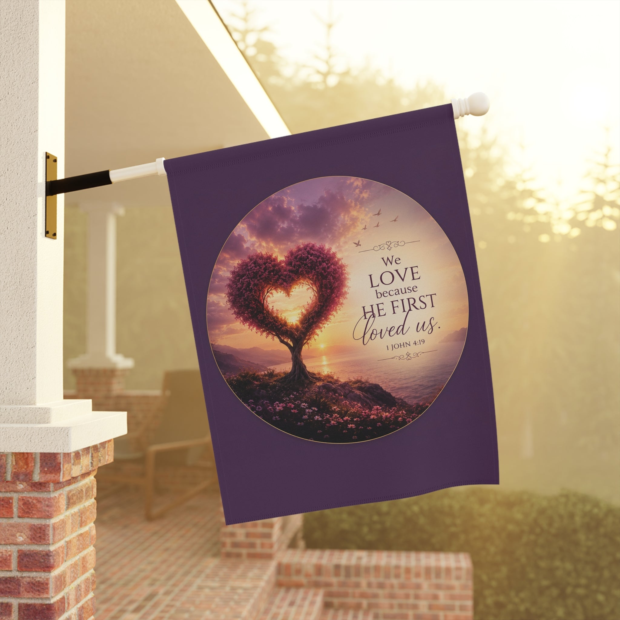 Inspirational Christian garden flag featuring a heart-shaped tree at sunset with the scripture “We Love Because He First Loved Us” in warm glowing tones.