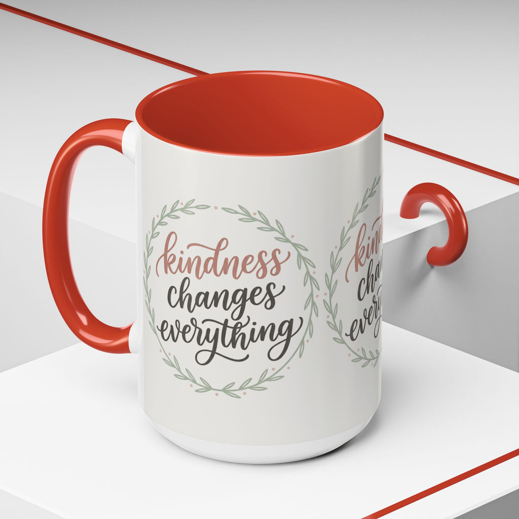 Inspirational ceramic accent coffee mug featuring hand-lettered “Kindness Changes Everything” typography surrounded by a delicate laurel wreath in soft sage and blush tones.