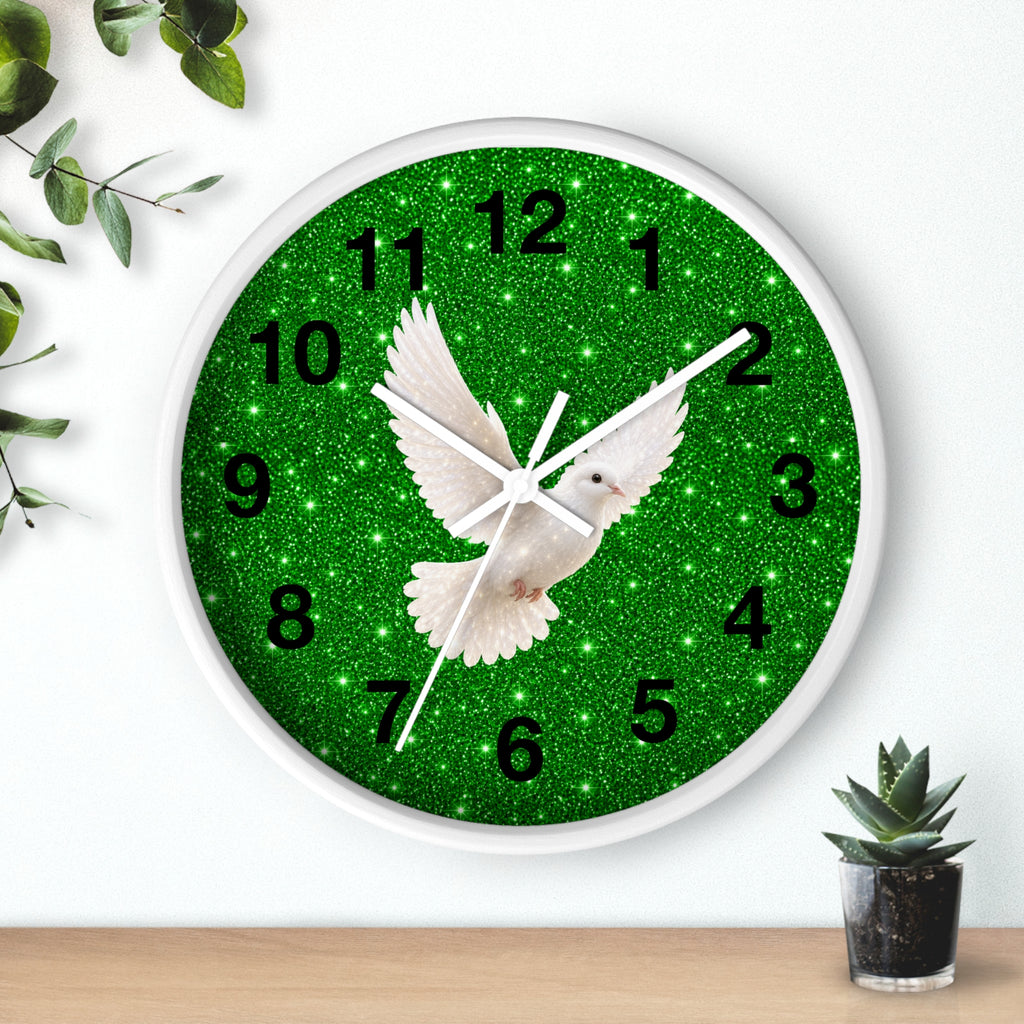 A round wall clock with a white frame and a green glitter-style face featuring a white dove in flight and black numerals, shown mounted on a white wall above a wooden surface with small plants nearby.