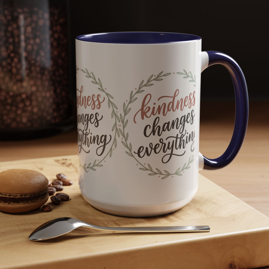Inspirational ceramic accent coffee mug featuring hand-lettered “Kindness Changes Everything” typography surrounded by a delicate laurel wreath in soft sage and blush tones.