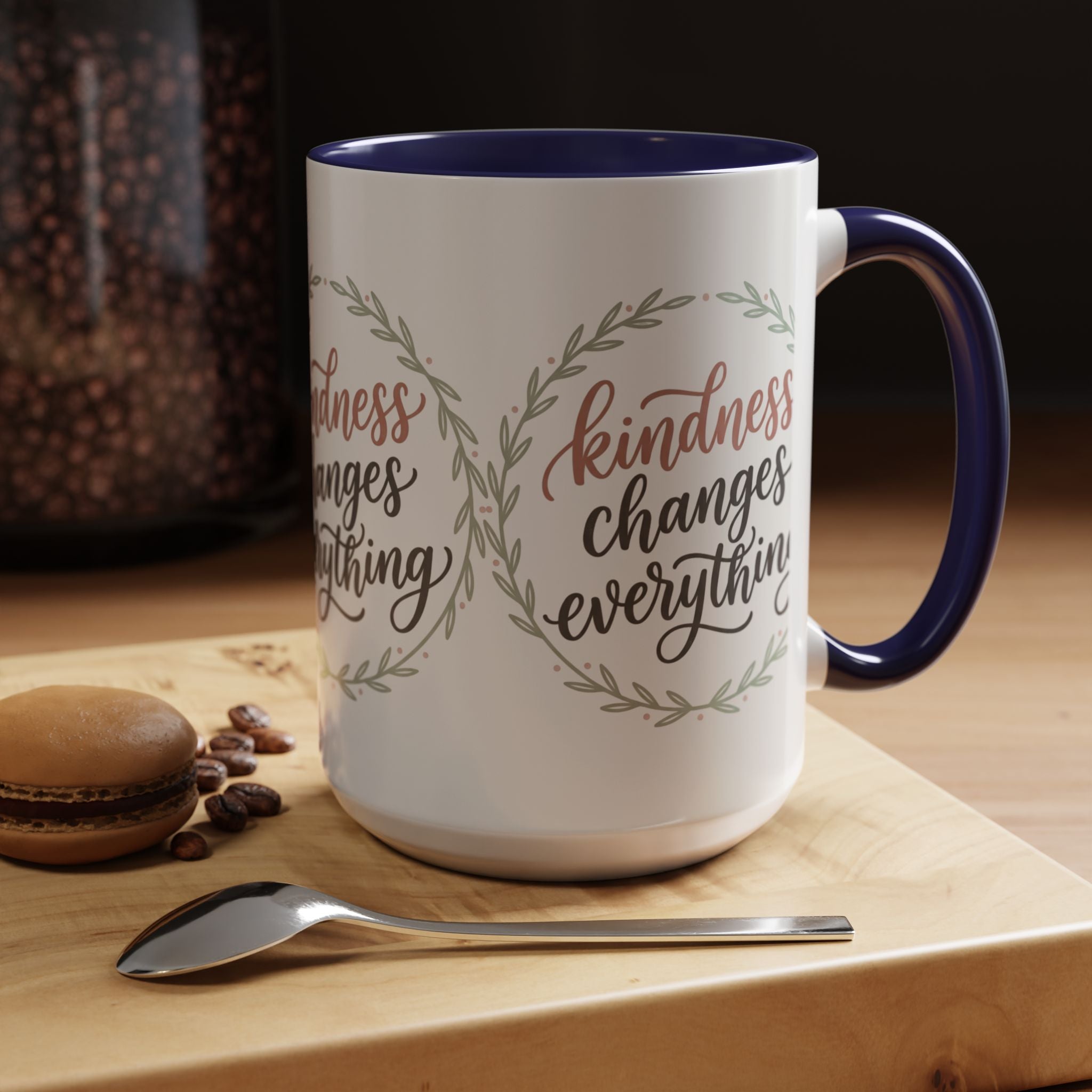 Inspirational ceramic accent coffee mug featuring hand-lettered “Kindness Changes Everything” typography surrounded by a delicate laurel wreath in soft sage and blush tones.