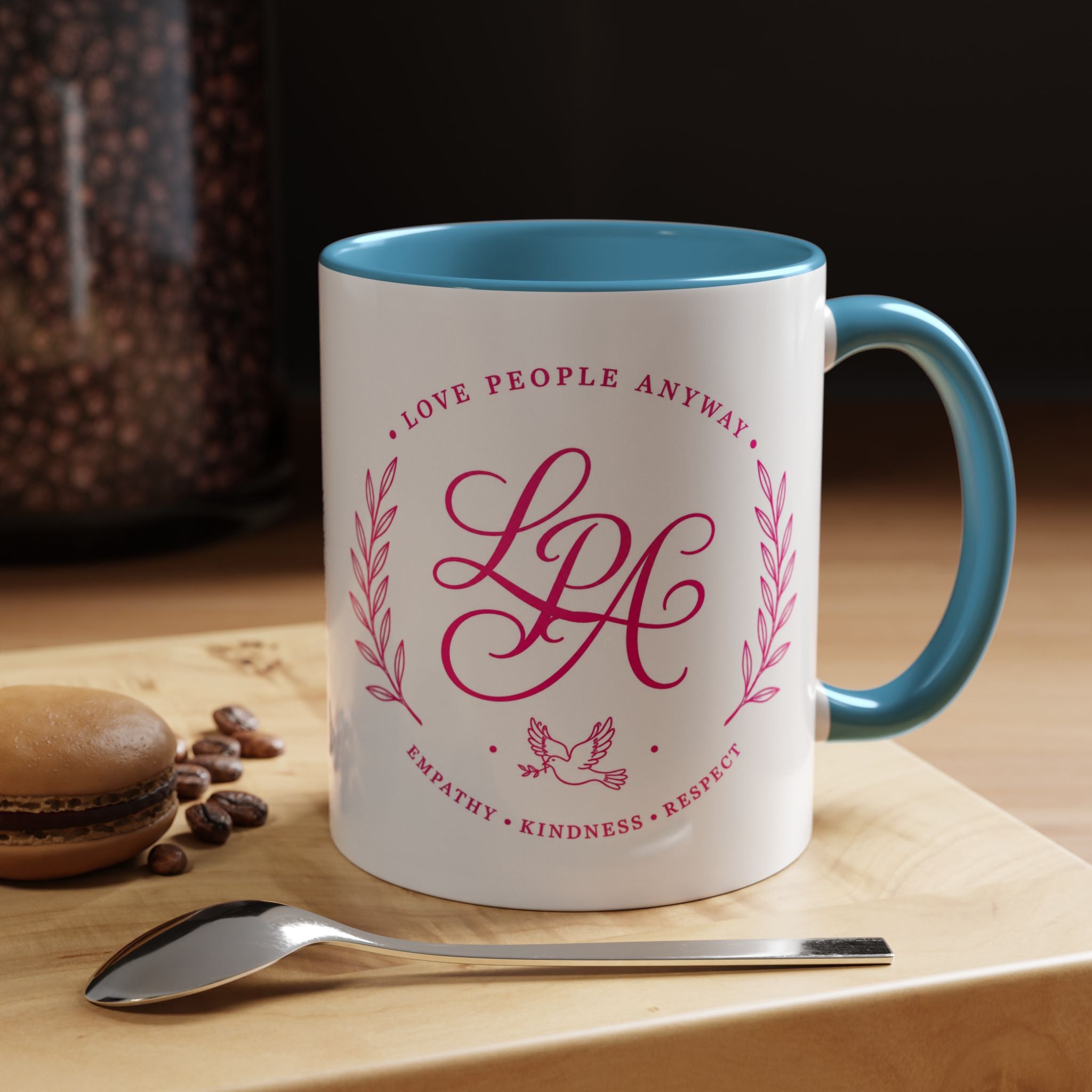 Inspirational ceramic coffee mug featuring soft pink “Love People Anyway” lettering, laurel accents, a dove motif, and contrasting light blue handle and interior.