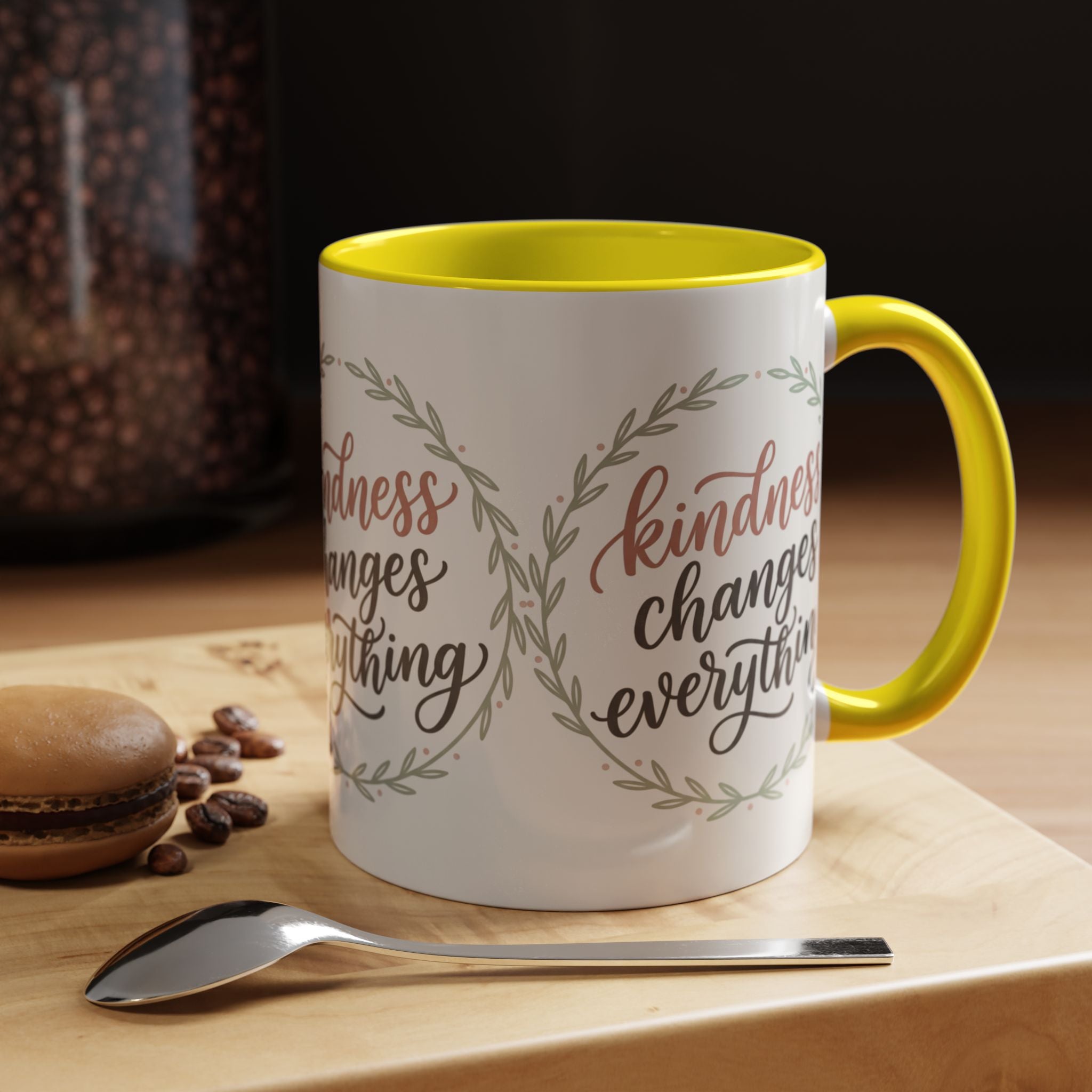 Inspirational ceramic accent coffee mug featuring hand-lettered “Kindness Changes Everything” typography surrounded by a delicate laurel wreath in soft sage and blush tones.