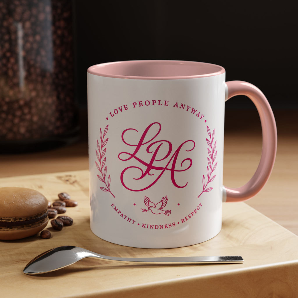 Inspirational ceramic coffee mug featuring soft pink “Love People Anyway” lettering, laurel accents, a dove motif, and contrasting black handle and interior.