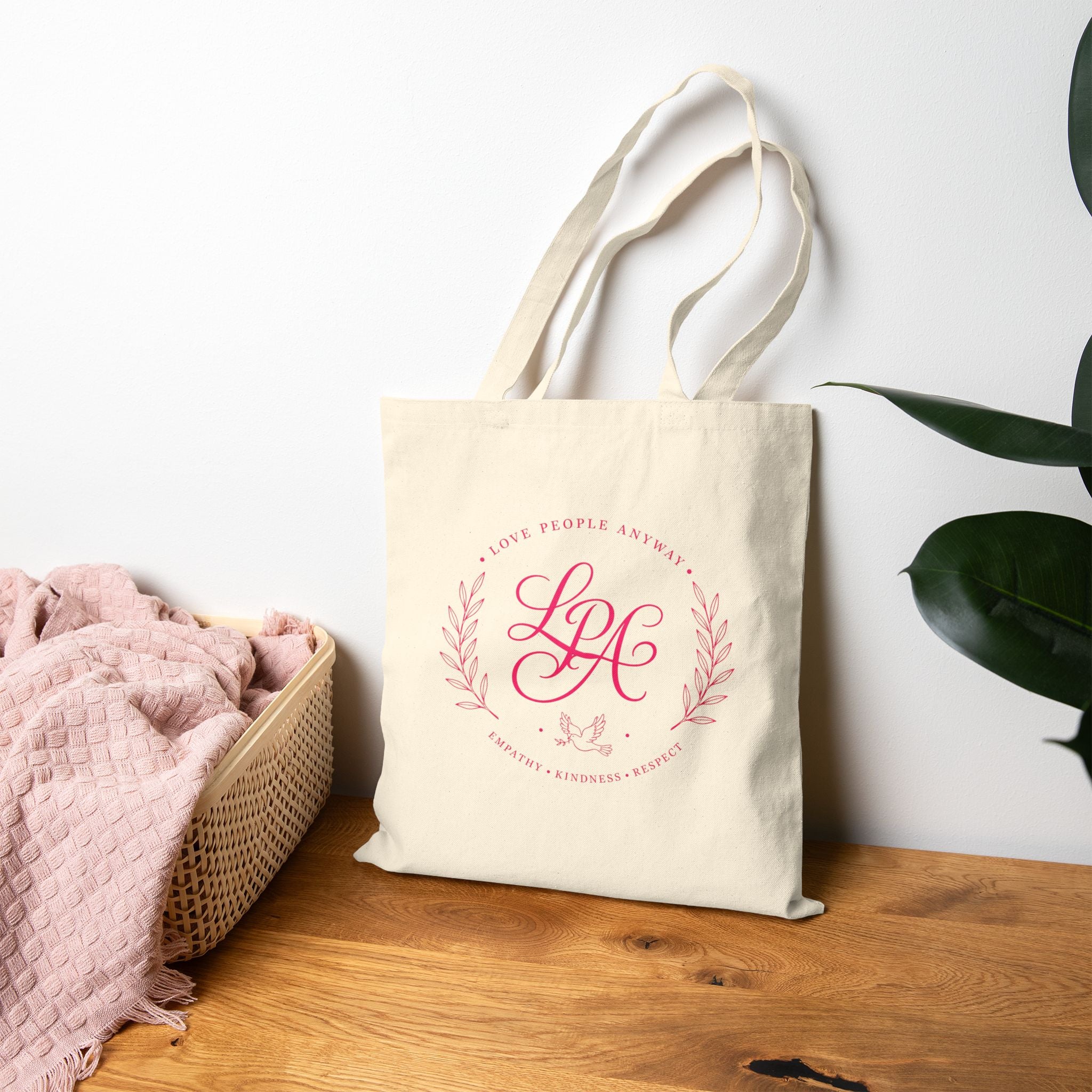 Pink monogram cotton canvas tote bag featuring the message “Love People Anyway,” laurel branches, a dove carrying an olive branch, and the words empathy, kindness, and respect in an elegant boutique design.