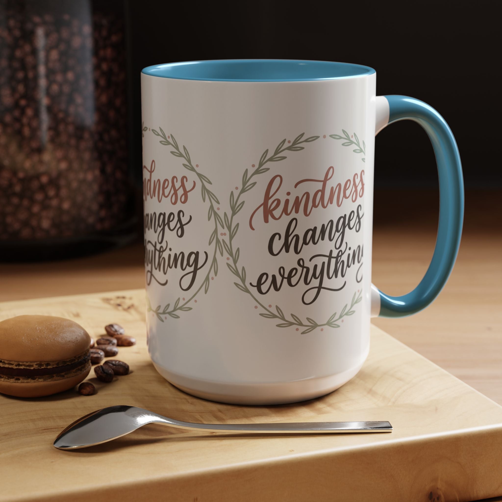 Inspirational ceramic accent coffee mug featuring hand-lettered “Kindness Changes Everything” typography surrounded by a delicate laurel wreath in soft sage and blush tones.