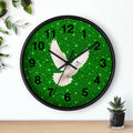 A round wall clock with a black frame and a green glitter face featuring a white dove in flight and black Arabic numerals, shown hanging on a white wall above a wooden surface with small plants.