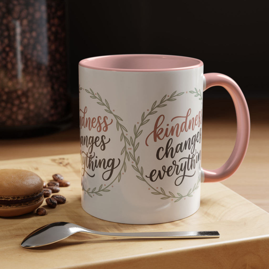 Inspirational ceramic accent coffee mug featuring hand-lettered “Kindness Changes Everything” typography surrounded by a delicate laurel wreath in soft sage and blush tones.