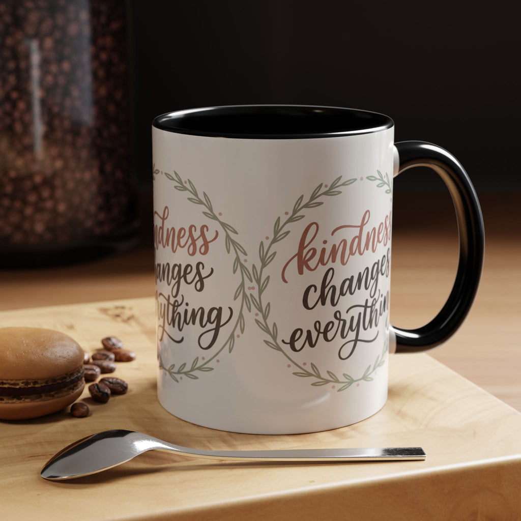 Inspirational ceramic accent coffee mug featuring hand-lettered “Kindness Changes Everything” typography surrounded by a delicate laurel wreath in soft sage and blush tones.