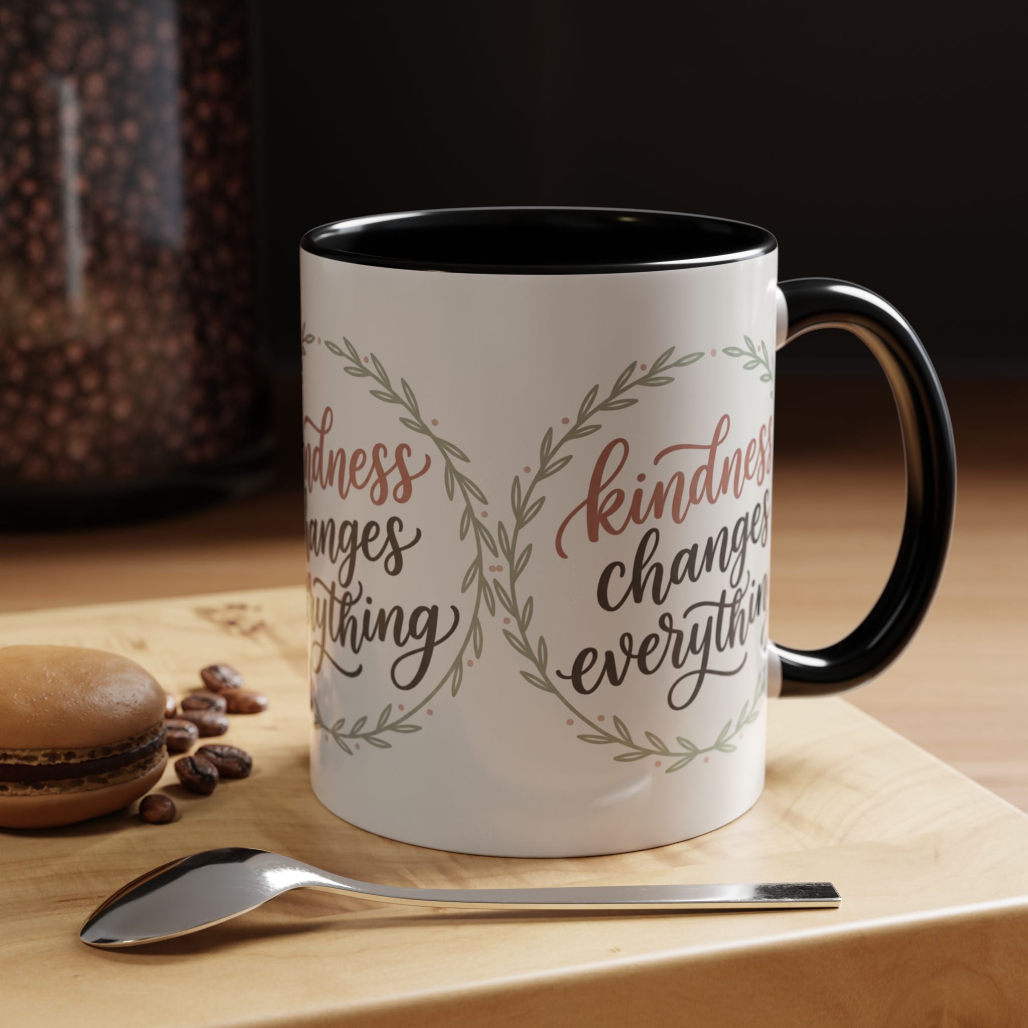 Inspirational ceramic accent coffee mug featuring hand-lettered “Kindness Changes Everything” typography surrounded by a delicate laurel wreath in soft sage and blush tones.