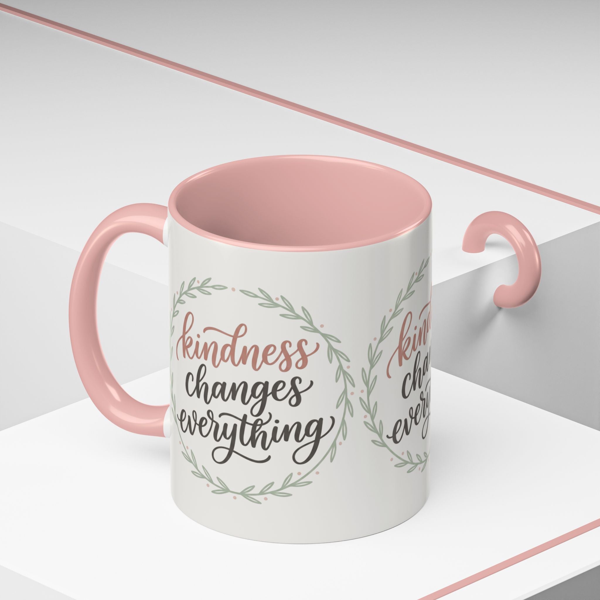 Inspirational ceramic accent coffee mug featuring hand-lettered “Kindness Changes Everything” typography surrounded by a delicate laurel wreath in soft sage and blush tones.