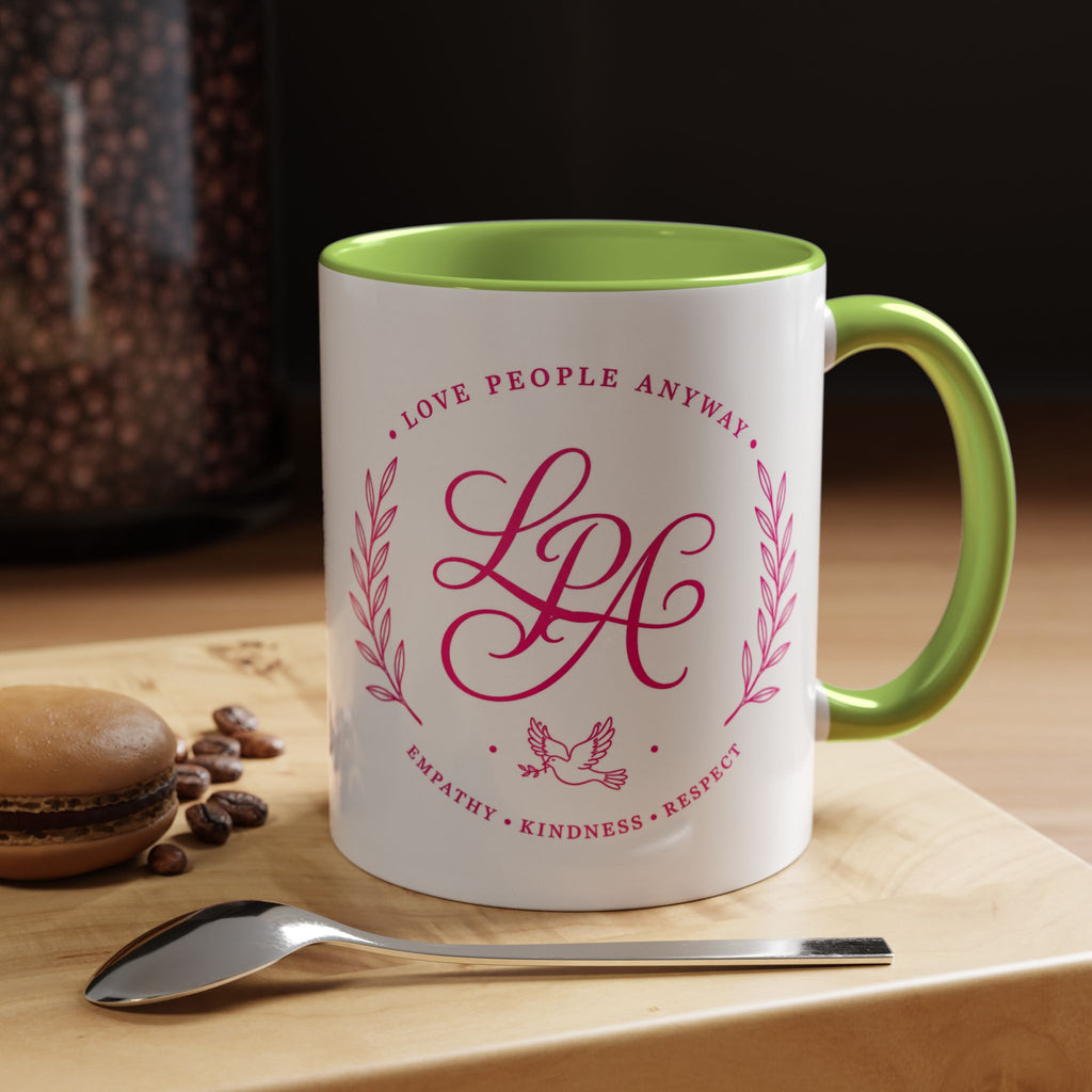 Inspirational ceramic coffee mug featuring soft pink “Love People Anyway” lettering, laurel accents, a dove motif, and contrasting green handle and interior.