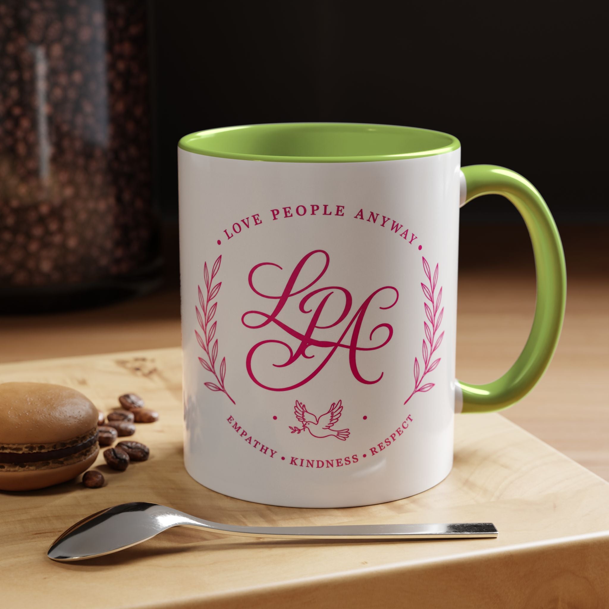 Inspirational ceramic coffee mug featuring soft pink “Love People Anyway” lettering, laurel accents, a dove motif, and contrasting green handle and interior.