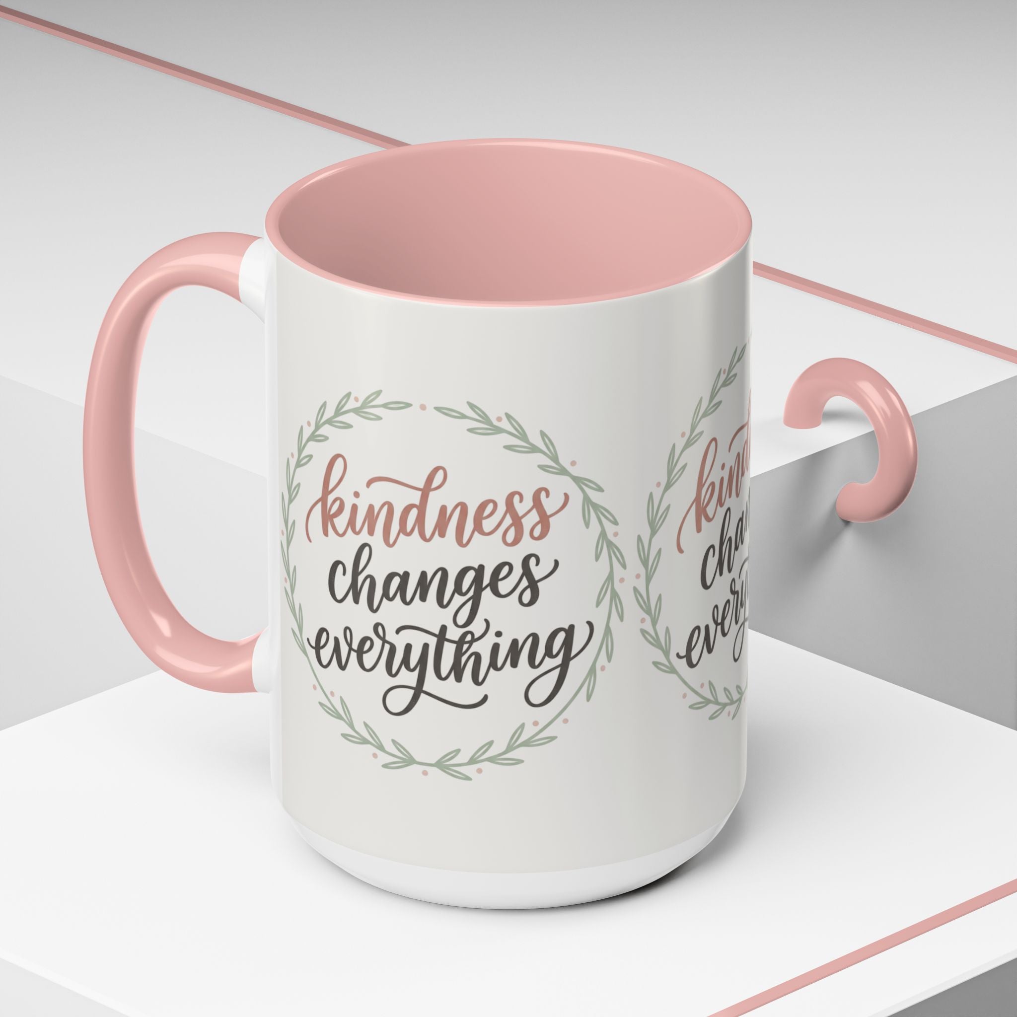 Inspirational ceramic accent coffee mug featuring hand-lettered “Kindness Changes Everything” typography surrounded by a delicate laurel wreath in soft sage and blush tones.