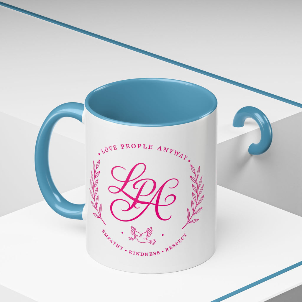 Inspirational ceramic coffee mug featuring soft pink “Love People Anyway” lettering, laurel accents, a dove motif, and contrasting light blue handle and interior.