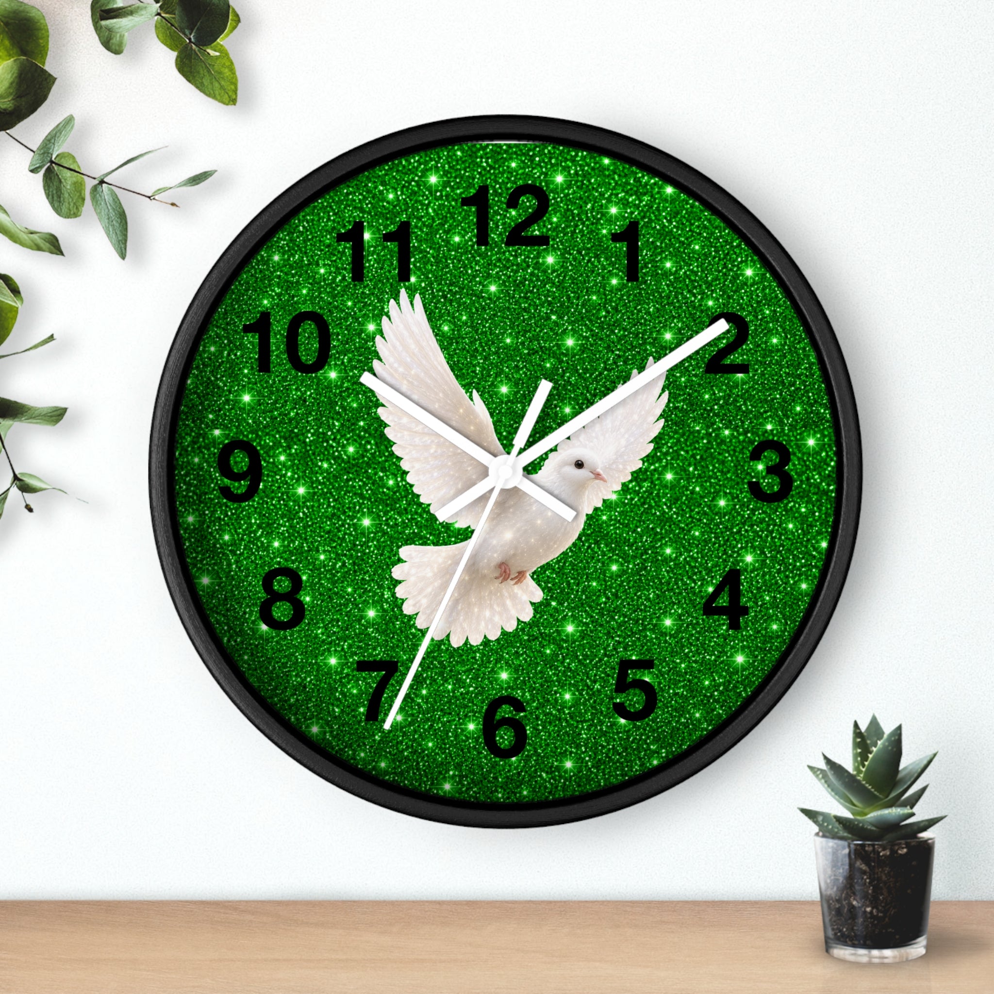 A round wall clock with a black frame and a green glitter face featuring a white dove in flight and black numerals, shown mounted on a white wall above a wooden surface with small plants.