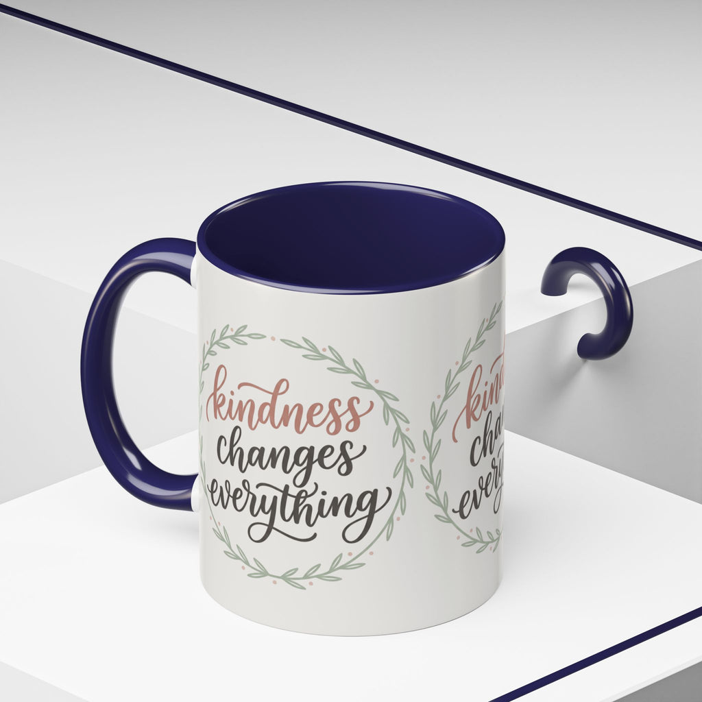 Inspirational ceramic accent coffee mug featuring hand-lettered “Kindness Changes Everything” typography surrounded by a delicate laurel wreath in soft sage and blush tones.