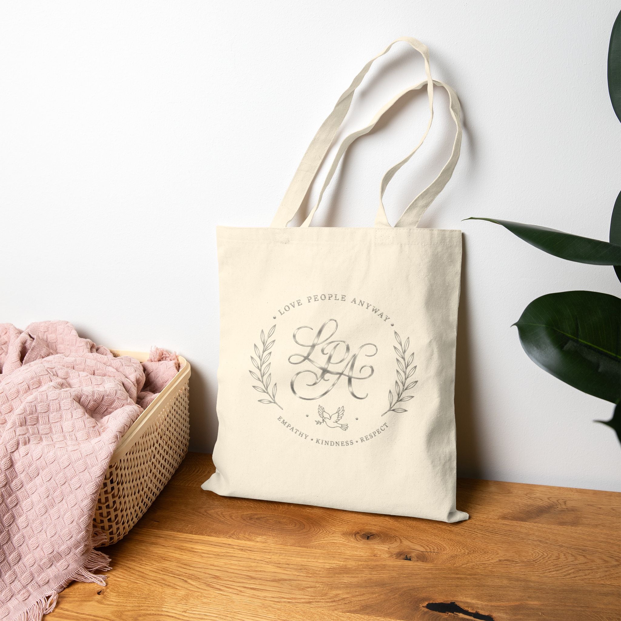 Elegant cotton canvas tote bag featuring a silver monogram crest, laurel branches, dove with olive branch, and the message “Love People Anyway” with empathy, kindness, and respect accents.