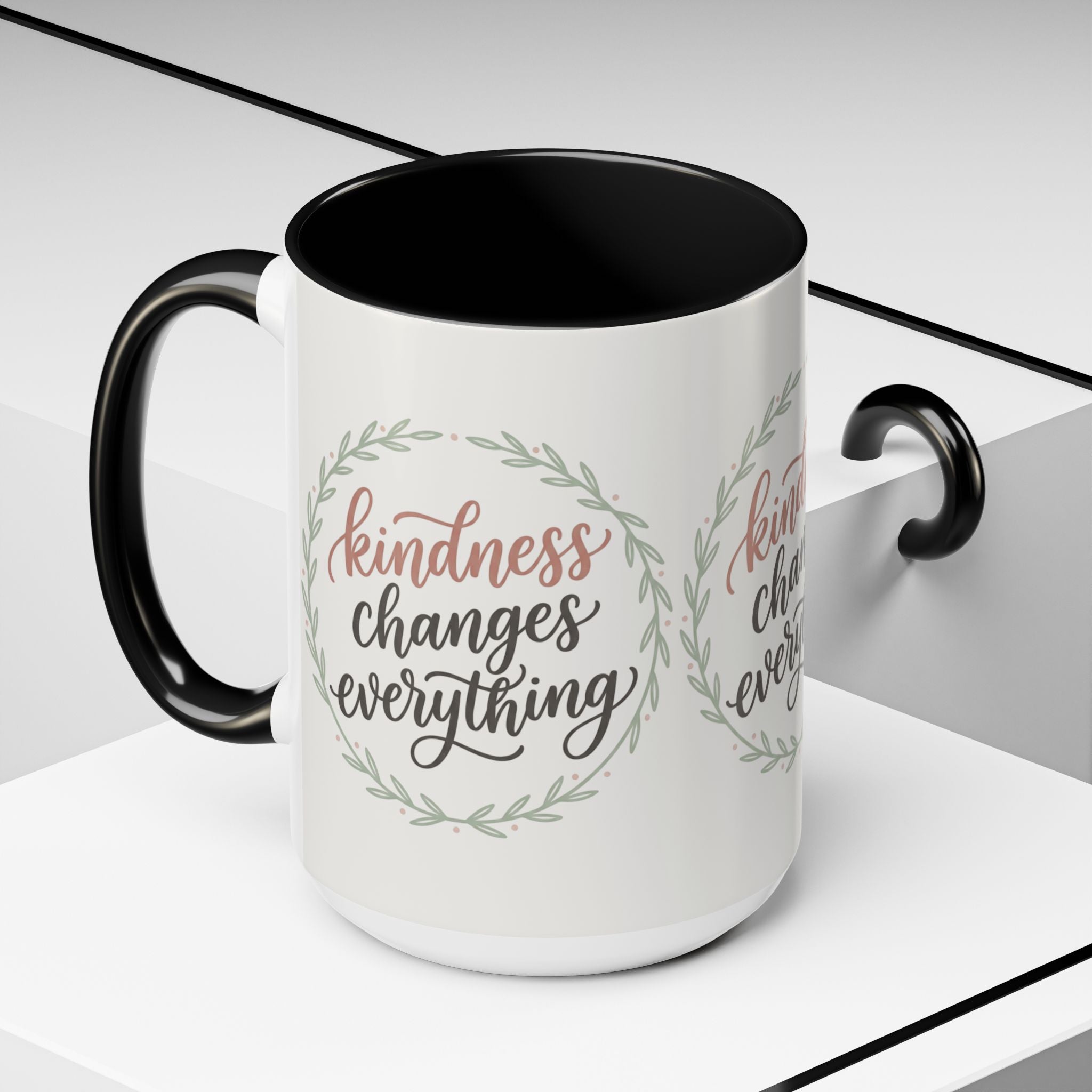 Inspirational ceramic accent coffee mug featuring hand-lettered “Kindness Changes Everything” typography surrounded by a delicate laurel wreath in soft sage and blush tones.