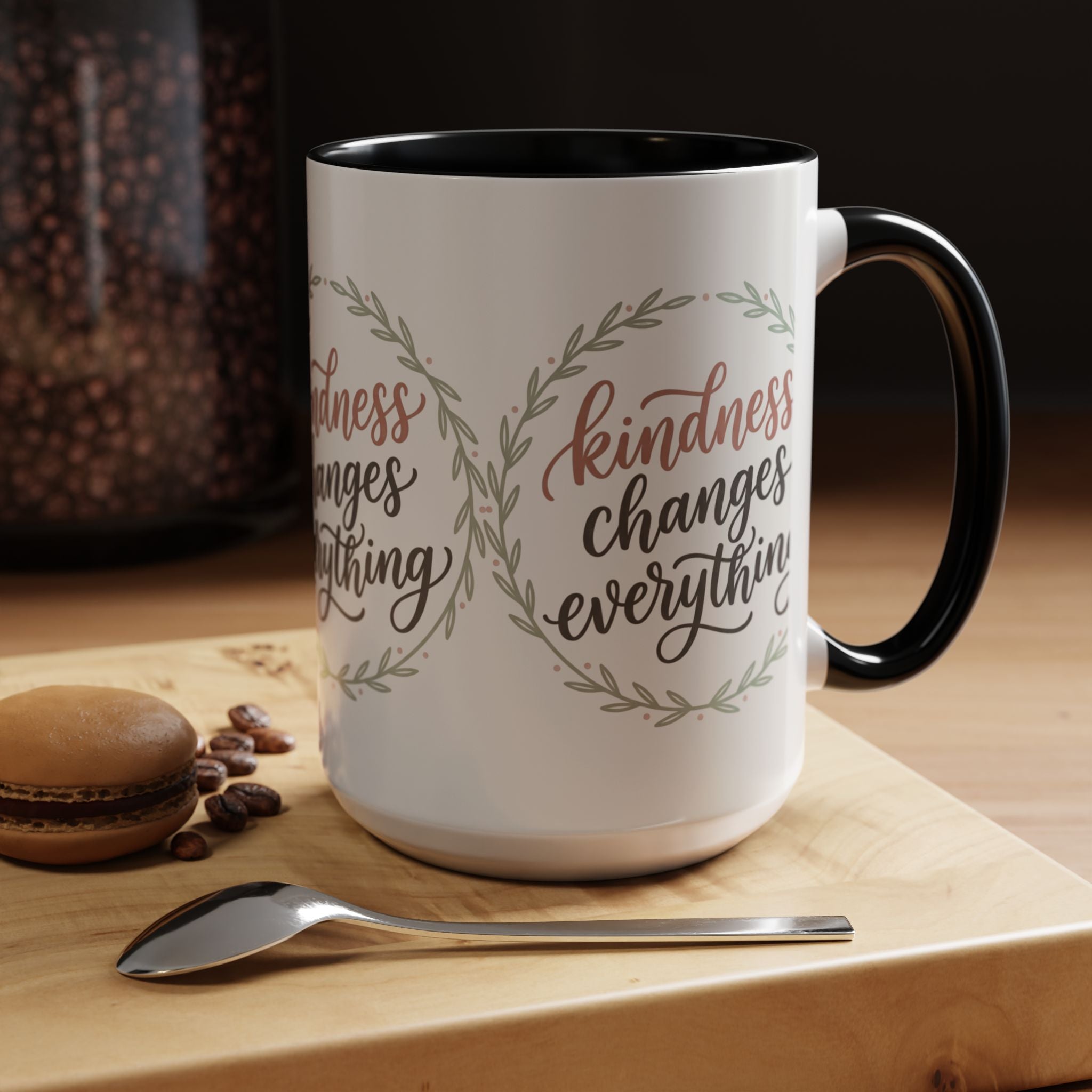 Inspirational ceramic accent coffee mug featuring hand-lettered “Kindness Changes Everything” typography surrounded by a delicate laurel wreath in soft sage and blush tones.