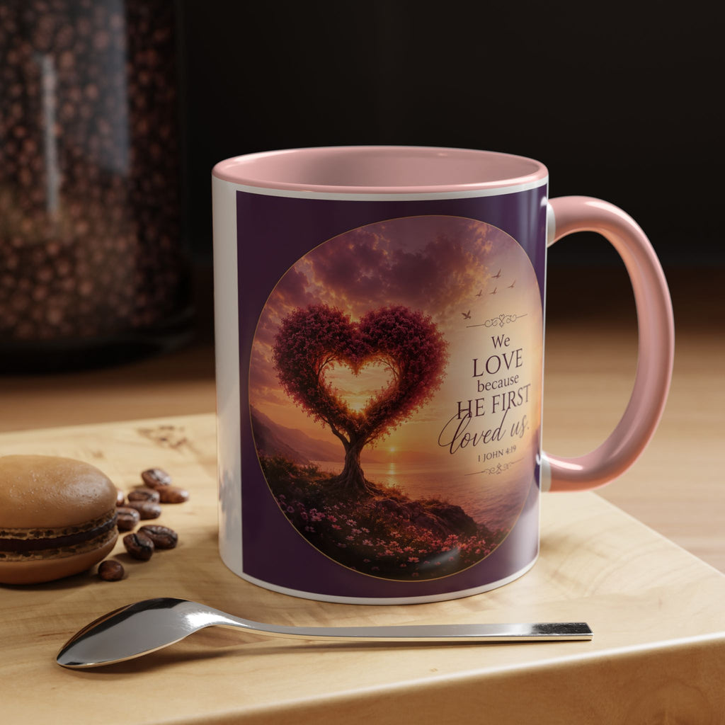Inspirational Christian coffee mug featuring a glowing heart-shaped tree at sunset with the scripture “We Love Because He First Loved Us” from 1 John 4:19 and contrasting black handle and interior.