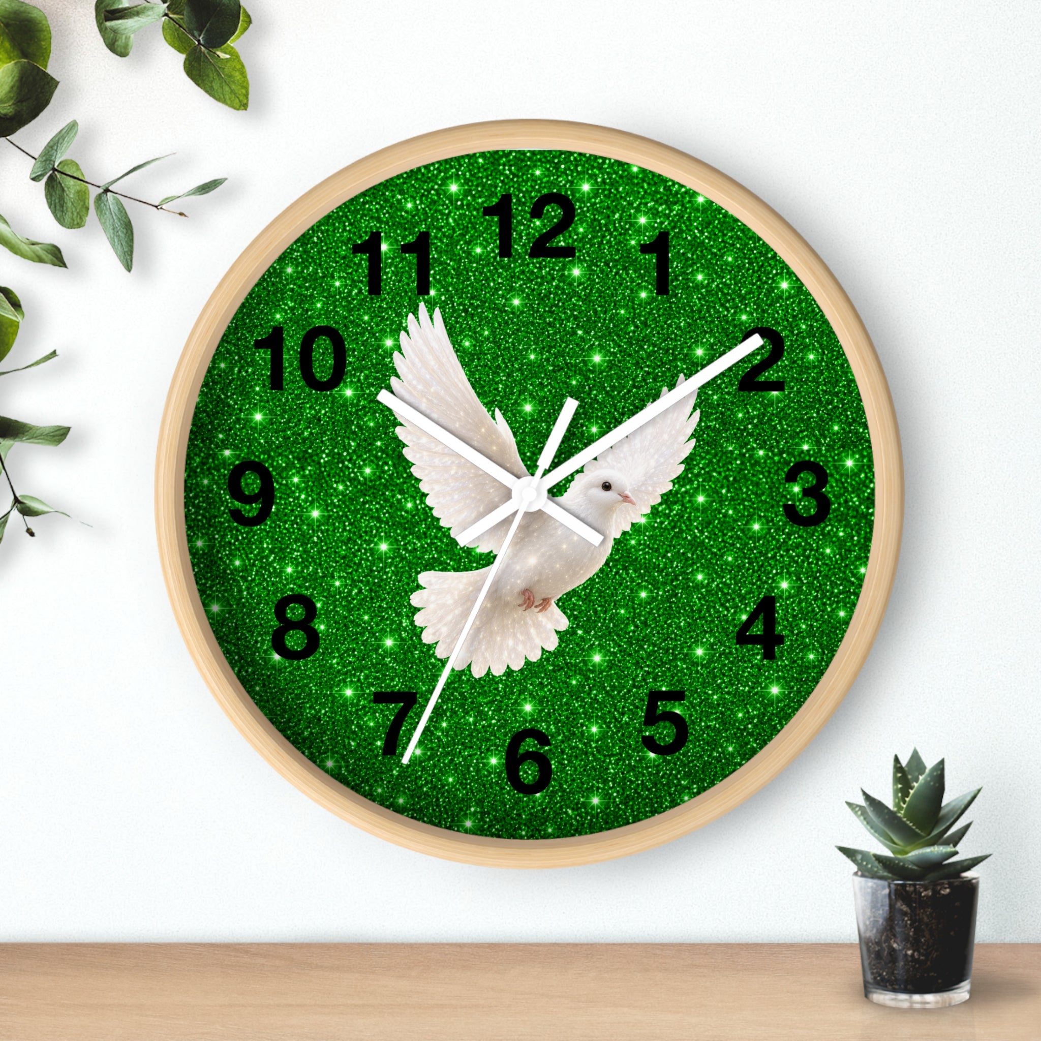 A round wall clock with a light wood frame, featuring a green glitter background, black Arabic numerals, and a white dove in flight at the center. The clock is shown hanging on a white wall above a wooden shelf with small decorative plants.