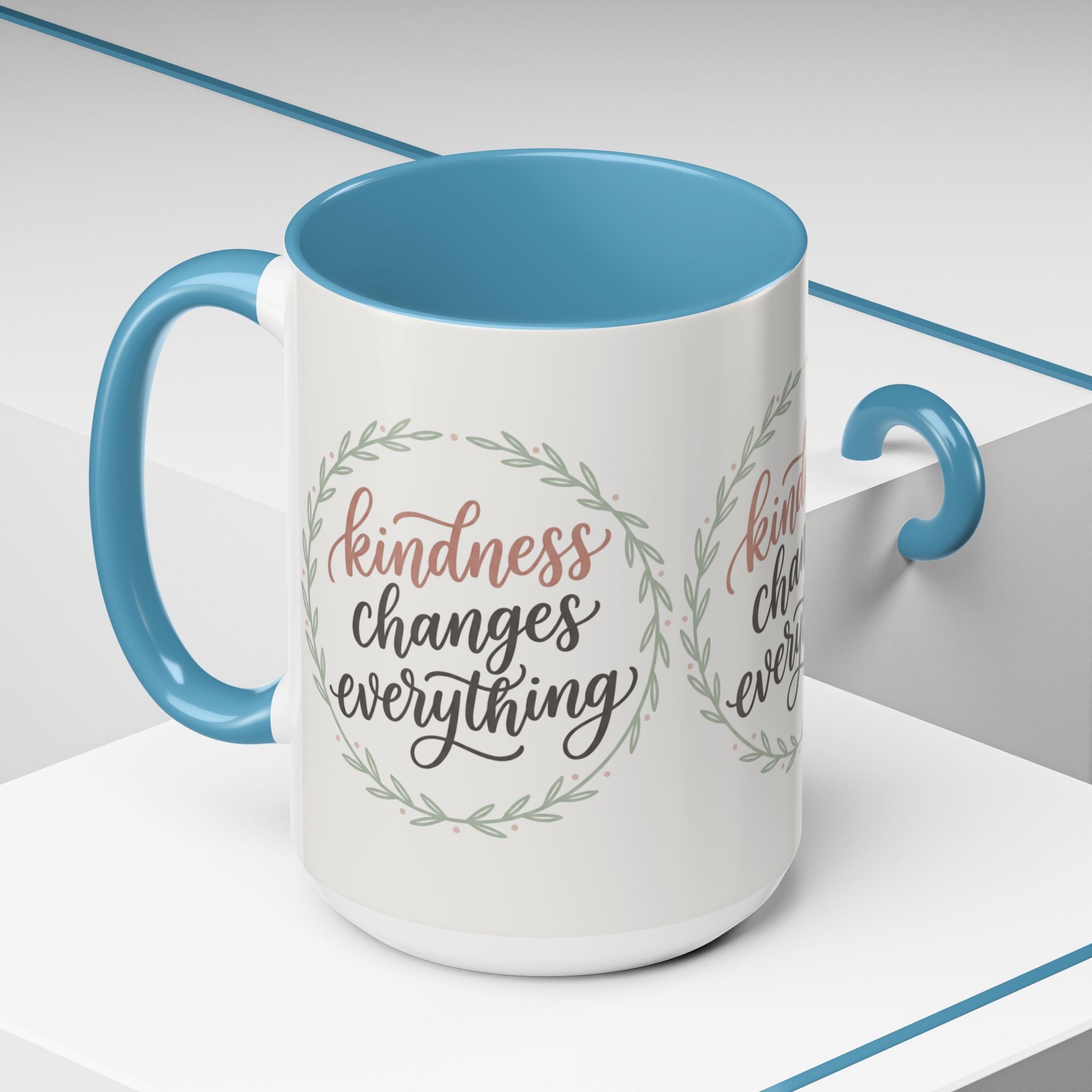 Inspirational ceramic accent coffee mug featuring hand-lettered “Kindness Changes Everything” typography surrounded by a delicate laurel wreath in soft sage and blush tones.