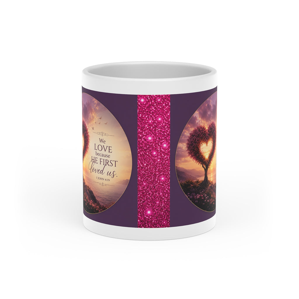 White ceramic heart-shaped handle mug featuring a glowing heart-shaped tree at sunset with the Scripture "We Love Because He First Loved Us" from 1 John 4:19 in rich jewel-toned artwork.