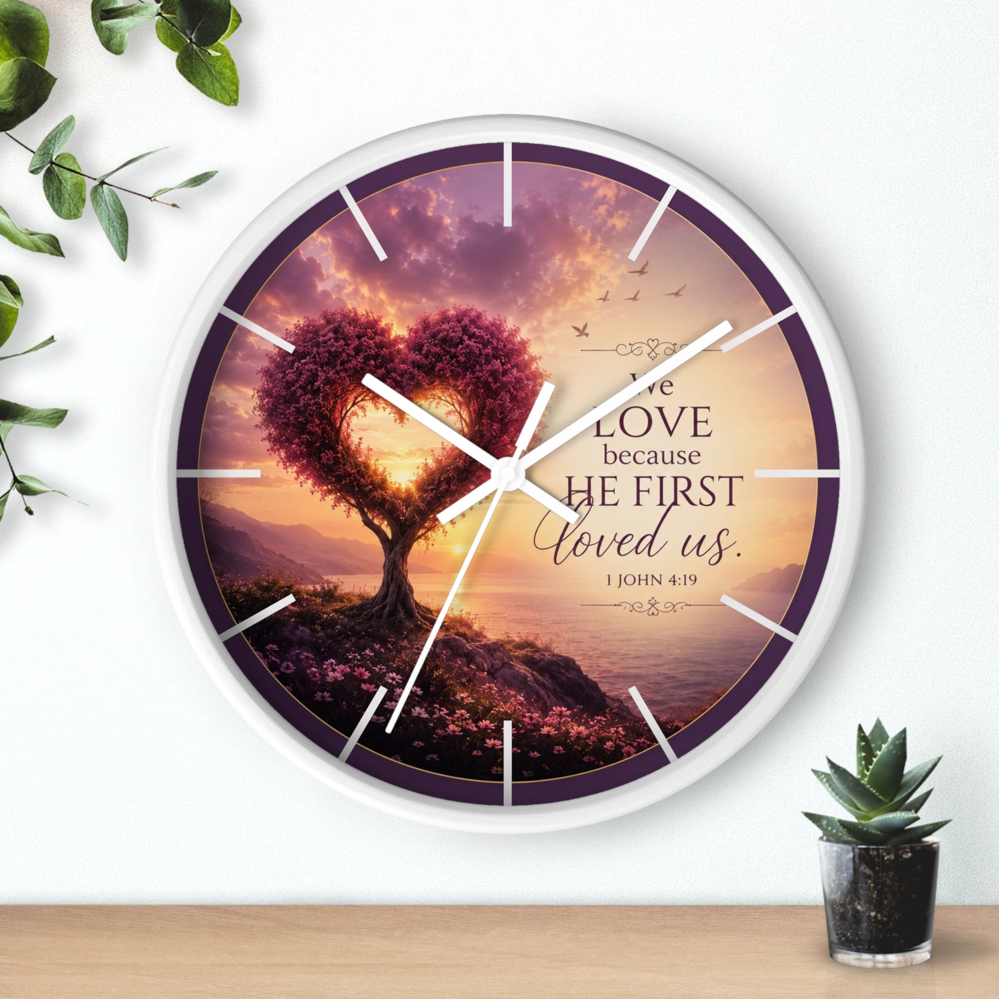 Inspirational Christian wall clock featuring a heart-shaped tree beside a peaceful shoreline sunset with 1 John 4:19 scripture about love and faith.