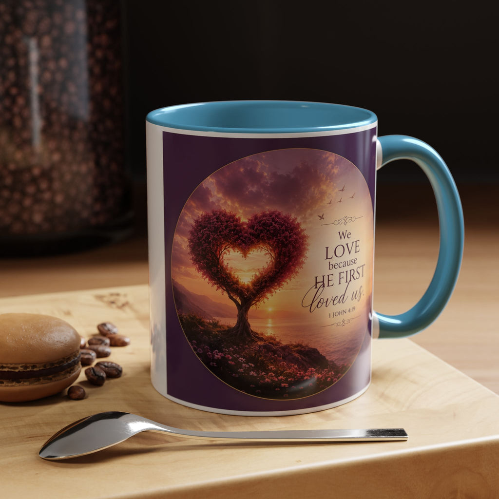 Inspirational Christian coffee mug featuring a glowing heart-shaped tree at sunset with the scripture “We Love Because He First Loved Us” from 1 John 4:19 and contrasting black handle and interior.