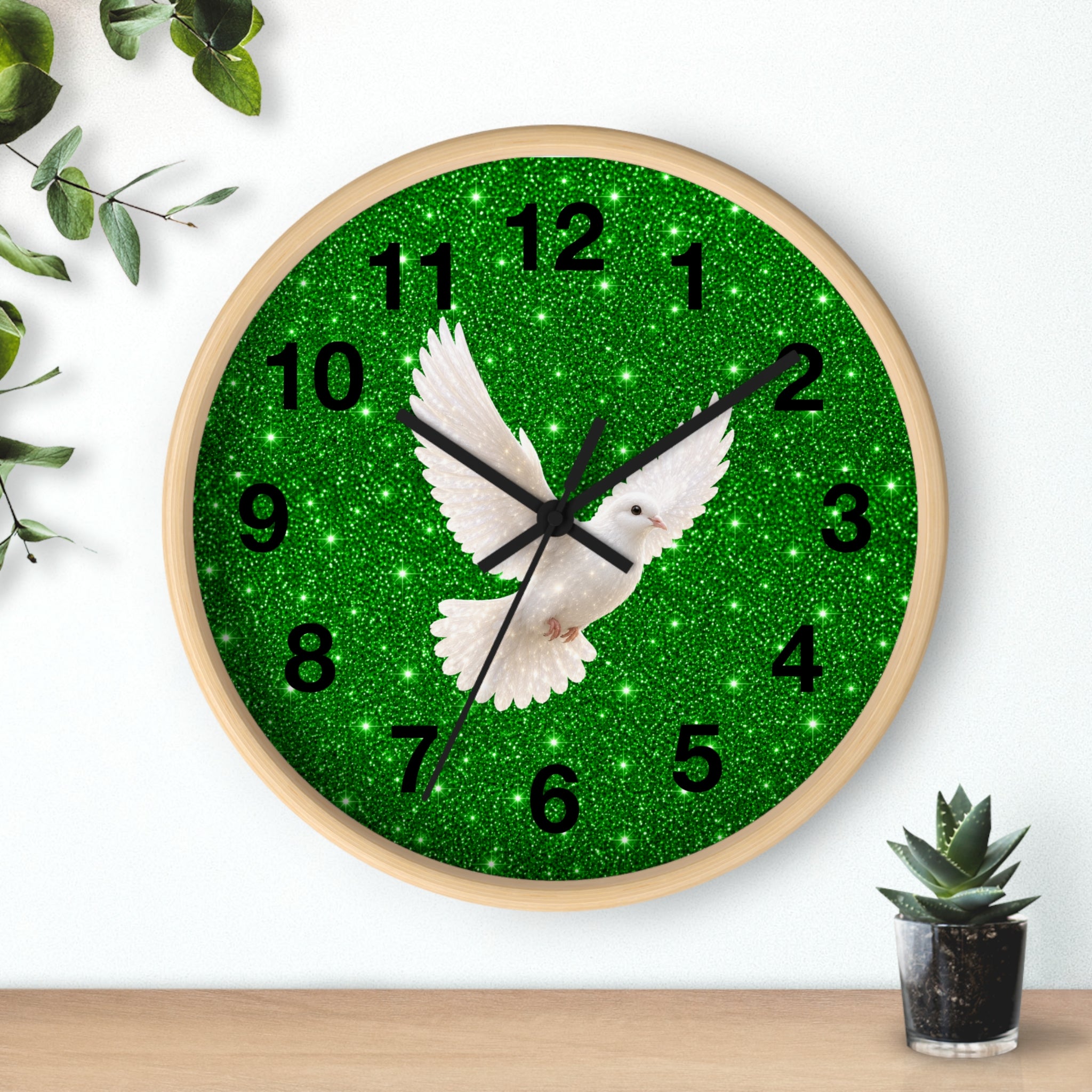 A round wall clock with a light wood frame, featuring a green glitter-textured face with black Arabic numerals and a white dove in flight at the center. The clock is mounted on a white wall above a light wood surface, with decorative green leaves on the left and a small potted succulent on the right.
