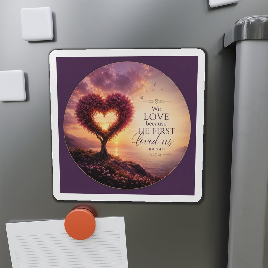 Die-cut Christian magnet featuring a heart-shaped tree at sunset with the scripture “We Love Because He First Loved Us” from 1 John 4:19 in purple, pink, and gold tones overlooking a peaceful shoreline.