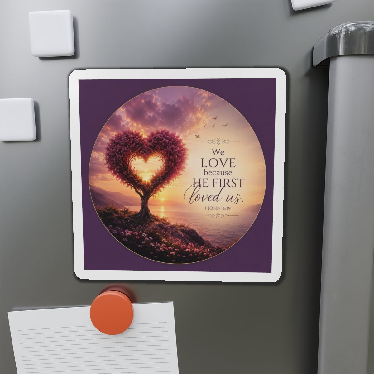Die-cut Christian magnet featuring a heart-shaped tree at sunset with the scripture “We Love Because He First Loved Us” from 1 John 4:19 in purple, pink, and gold tones overlooking a peaceful shoreline.