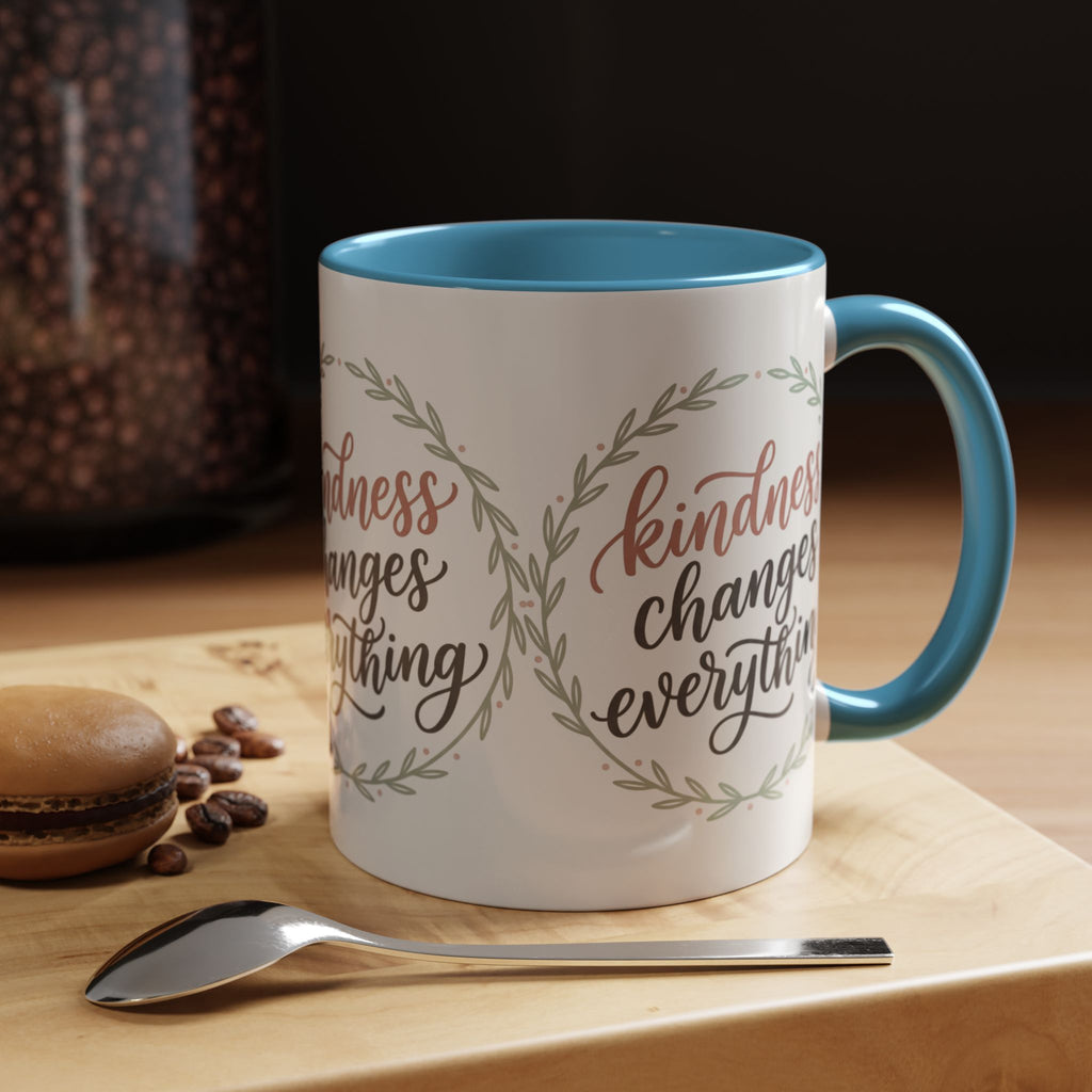 Inspirational ceramic accent coffee mug featuring hand-lettered “Kindness Changes Everything” typography surrounded by a delicate laurel wreath in soft sage and blush tones.