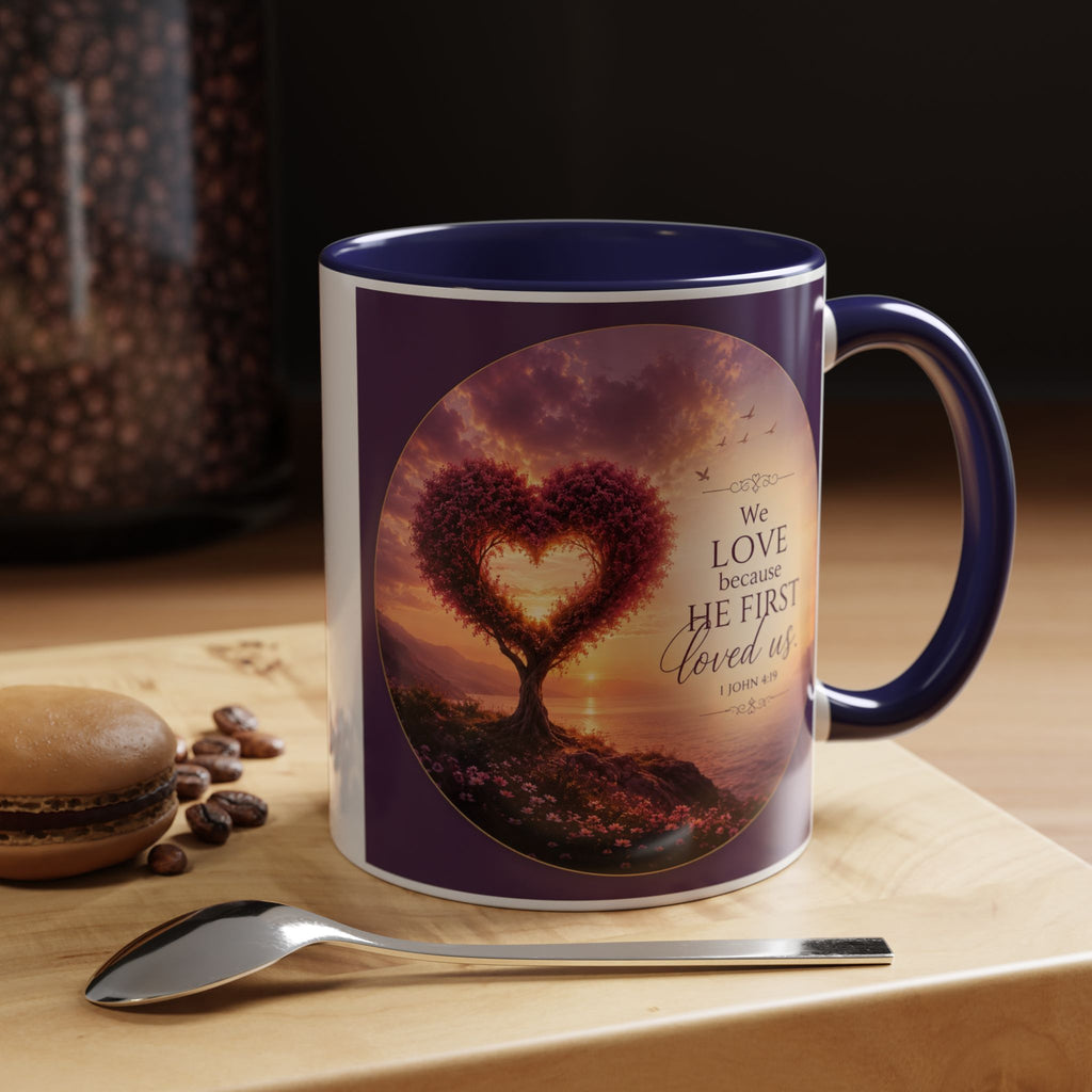 Inspirational Christian coffee mug featuring a glowing heart-shaped tree at sunset with the scripture “We Love Because He First Loved Us” from 1 John 4:19 and contrasting black handle and interior.