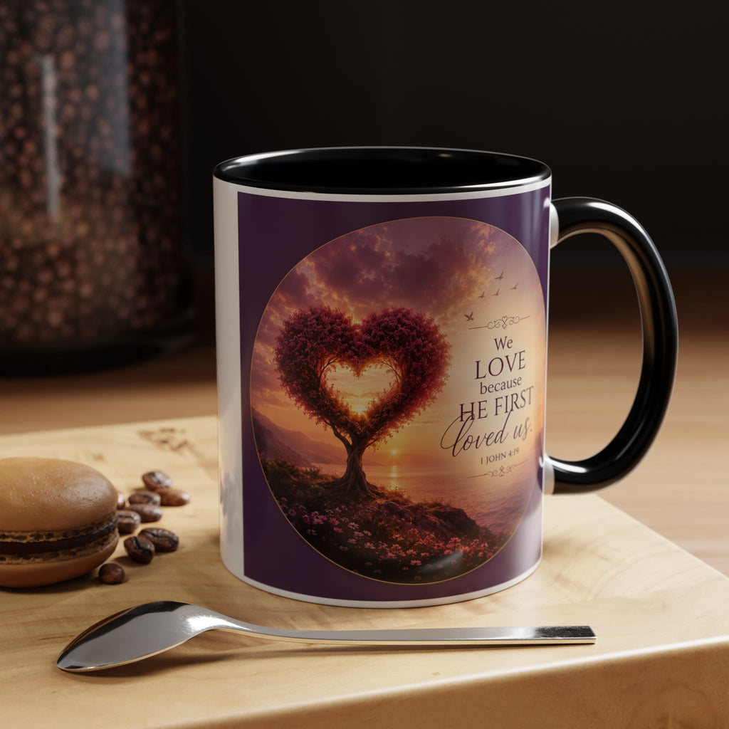Inspirational Christian coffee mug featuring a glowing heart-shaped tree at sunset with the scripture “We Love Because He First Loved Us” from 1 John 4:19 and contrasting black handle and interior.