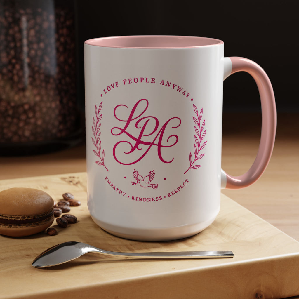 Inspirational ceramic coffee mug featuring soft pink “Love People Anyway” lettering, laurel accents, a dove motif, and contrasting pink handle and interior.