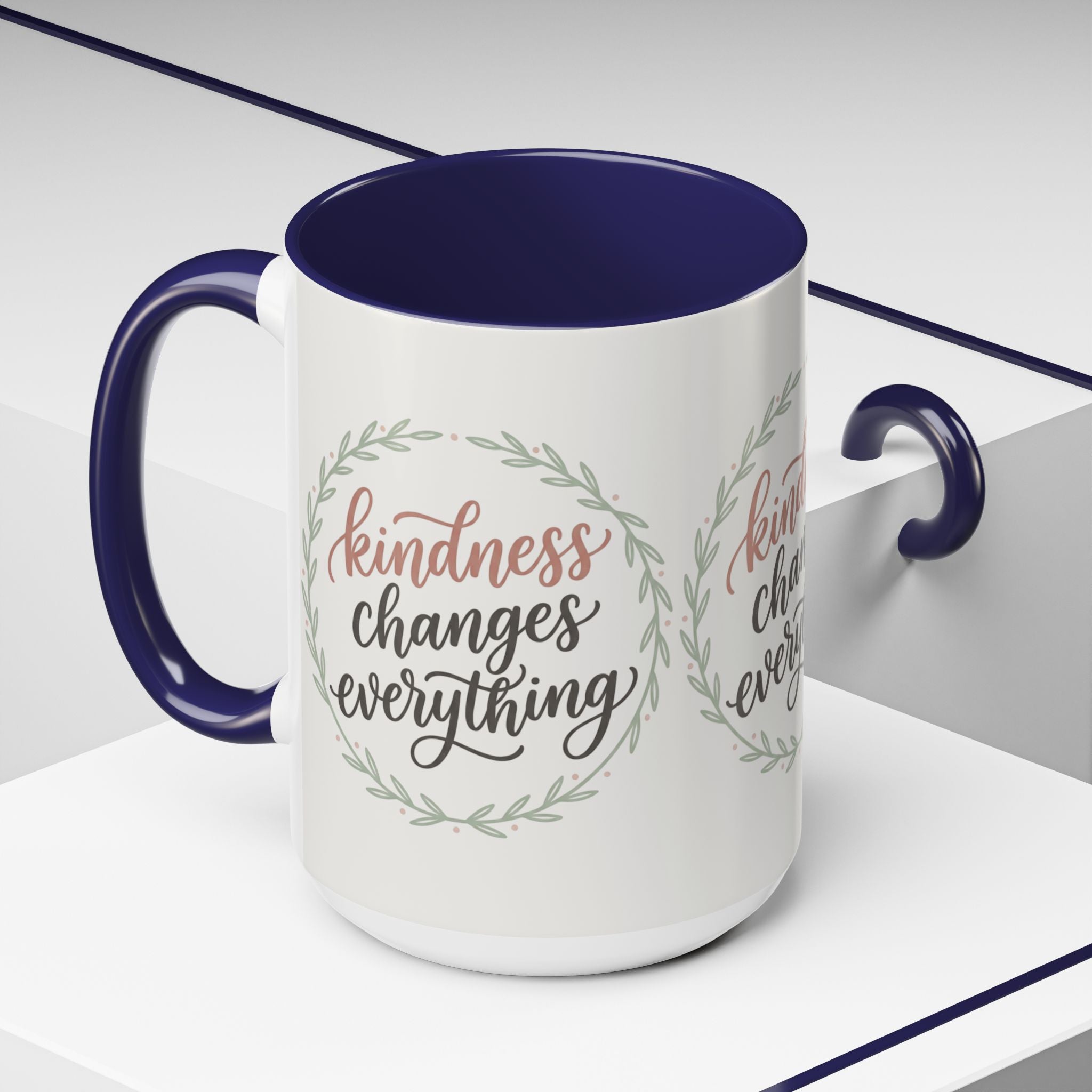 Inspirational ceramic accent coffee mug featuring hand-lettered “Kindness Changes Everything” typography surrounded by a delicate laurel wreath in soft sage and blush tones.