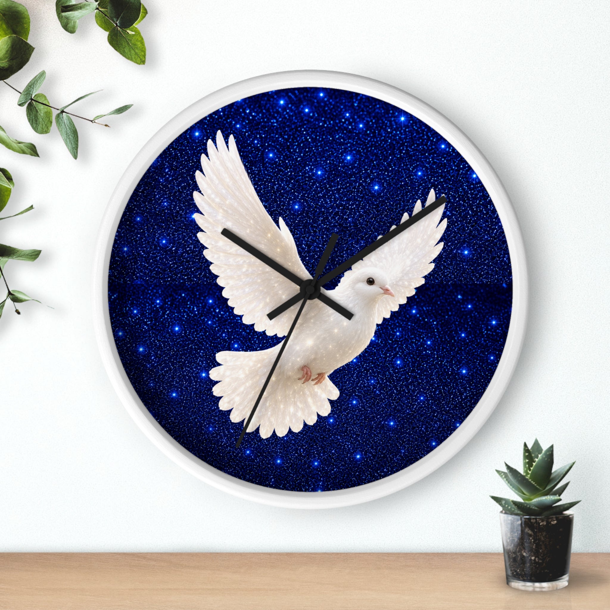Round wall clock with a white frame and a dark blue starry sky face, featuring a white dove in flight at the center. The clock is mounted on a white wall above a light wood surface, with decorative green leaves on the left and a small potted succulent on the right.